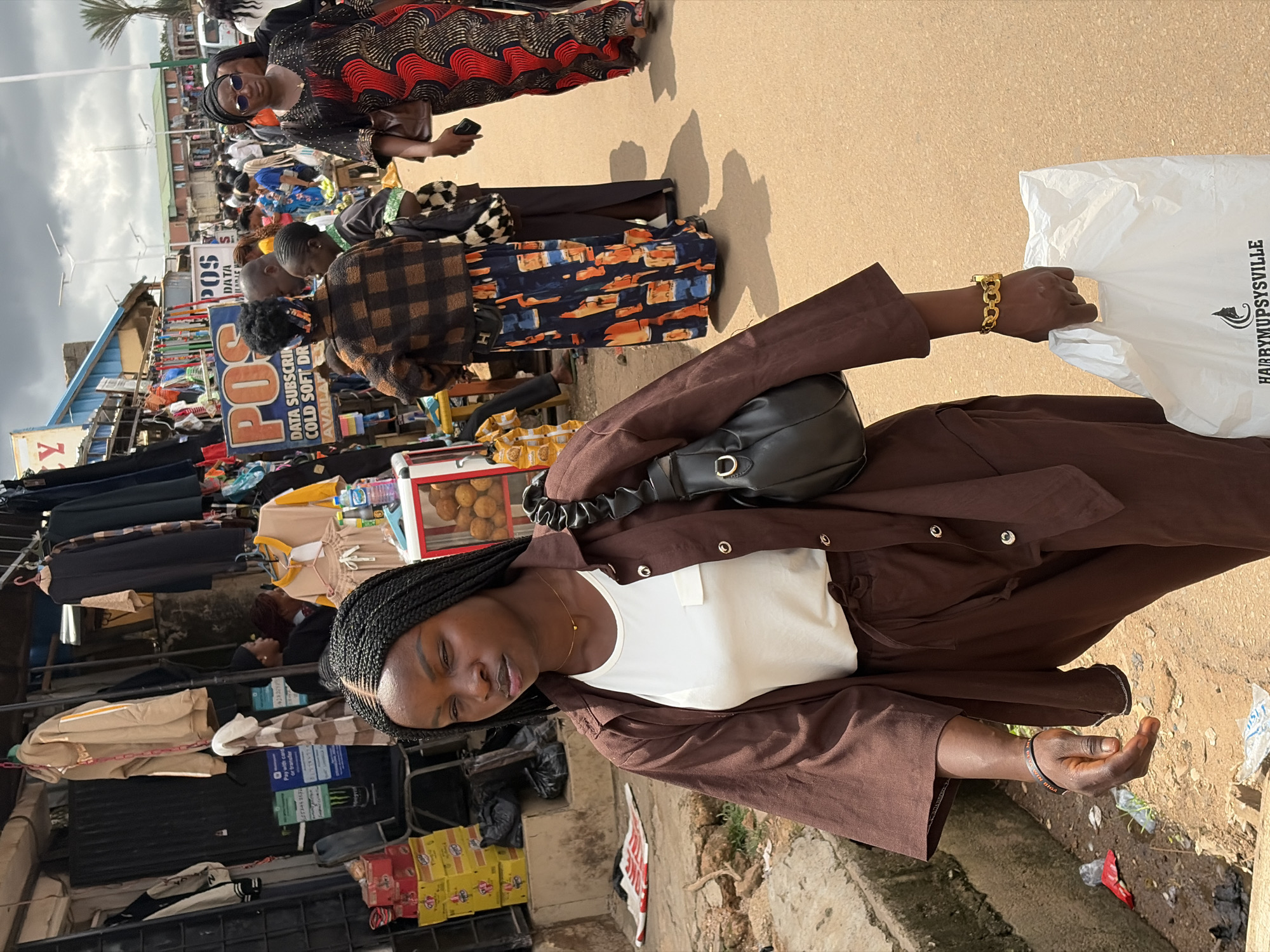 A young woman with braided hair, wearing a brown oversized button-up shirt over a white t-shirt and brown pants, walks forward in a market in Jos, Nigeria. She carries a black handbag on her left shoulder and a white plastic bag in her right hand. Behind her, other people, including women in patterned dresses, are browsing stalls laden with clothing and goods. The market appears to be outdoors with a cloudy sky. The ground is paved with reddish-brown dirt and concrete.