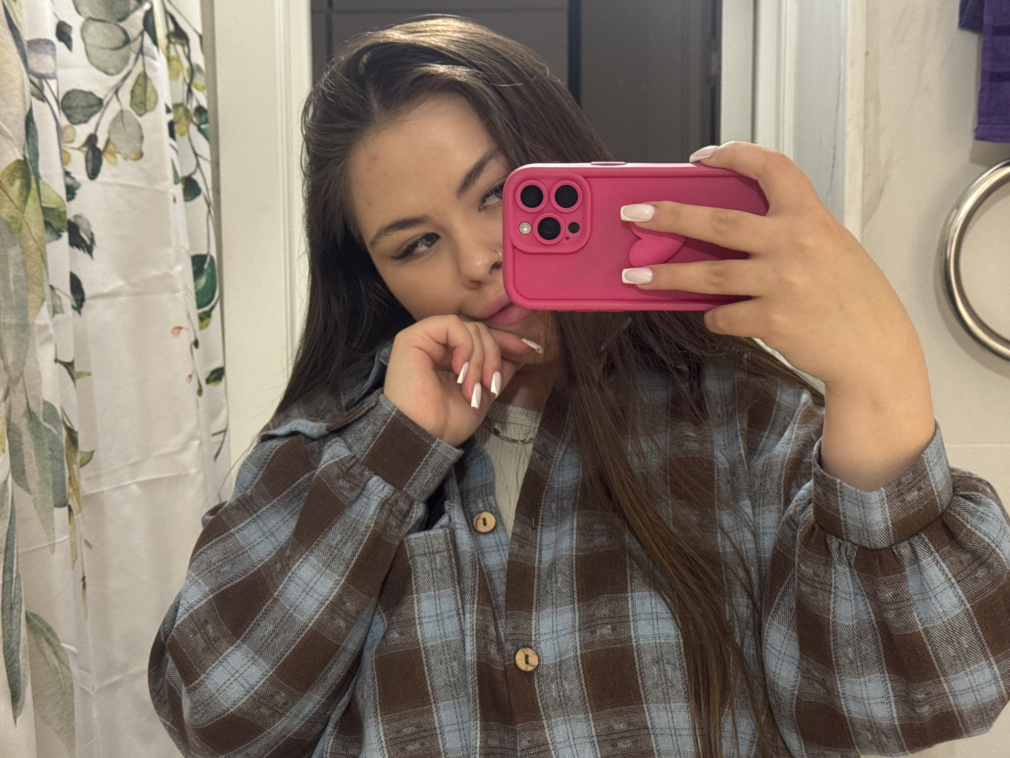 A young woman is taking a selfie in a bathroom. She is holding a pink smartphone with a heart-shaped detail on the back. She is wearing a plaid shirt with brown and light blue squares, and a light-colored shirt underneath. The bathroom features a shower curtain with a leaf pattern, a white door, and a towel rack. The location is Unknown, Unknown.