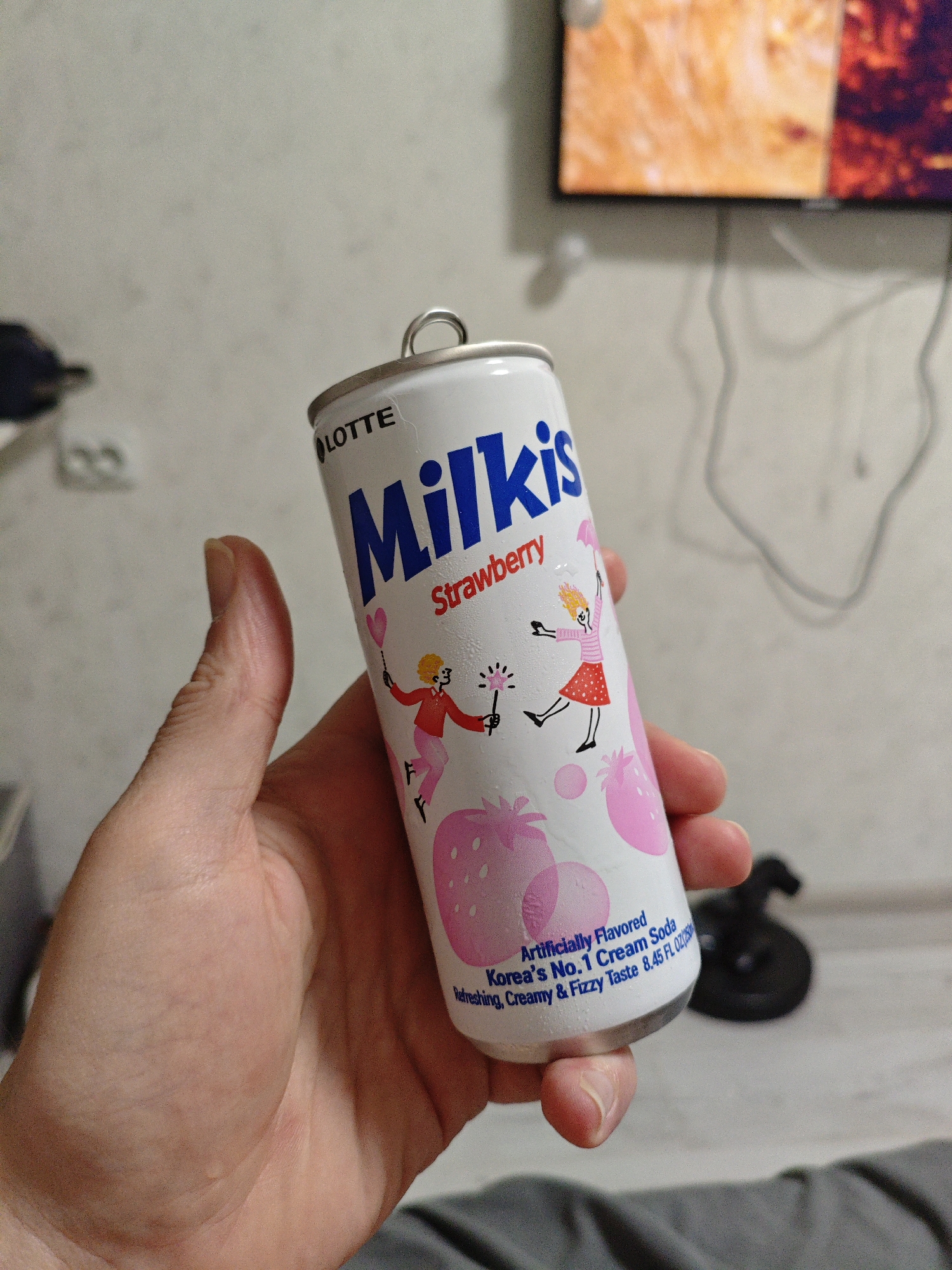 A person's hand holds a can of Lotte Milkis Strawberry cream soda. The can is white with blue lettering that reads "Milkis" and red lettering that says "Strawberry". The can also features cartoon drawings of people and strawberries. The background appears to be an indoor setting, possibly a room with a television screen visible in the upper right corner. The person's hand is light-skinned and the nails are short. The can is slightly damp with condensation. The image does not provide any information about the specific location within the city of Kyiv, Ukraine.