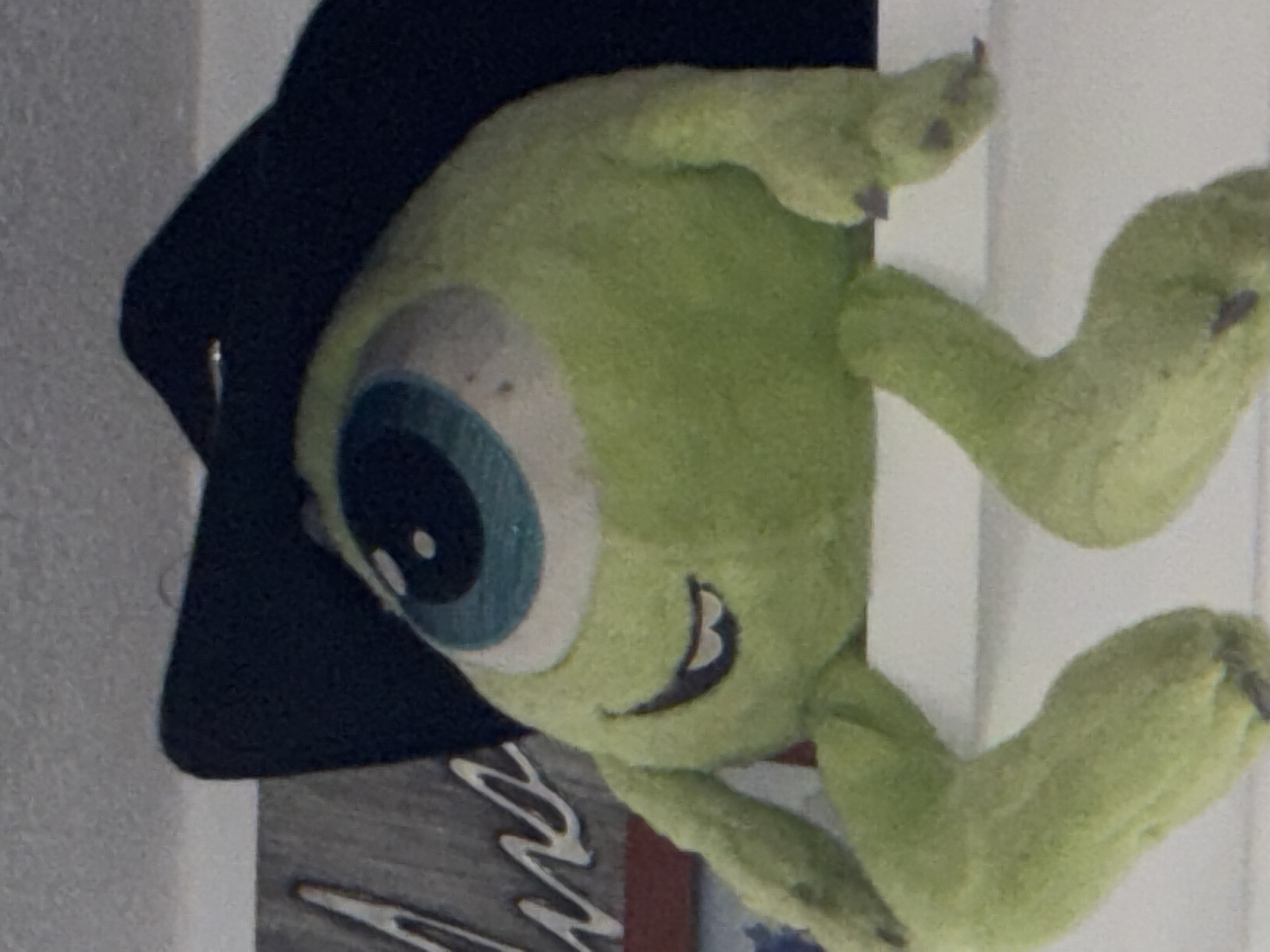 A plush, green, one-eyed toy resembling Mike Wazowski from "Monsters, Inc." wearing a black graduation cap is the main subject. The toy is situated against a predominantly white background, with a portion of a sign featuring gray and white abstract designs visible beneath it. The location is unknown, and there's no data to support it being Mexico City. No people are in the image.