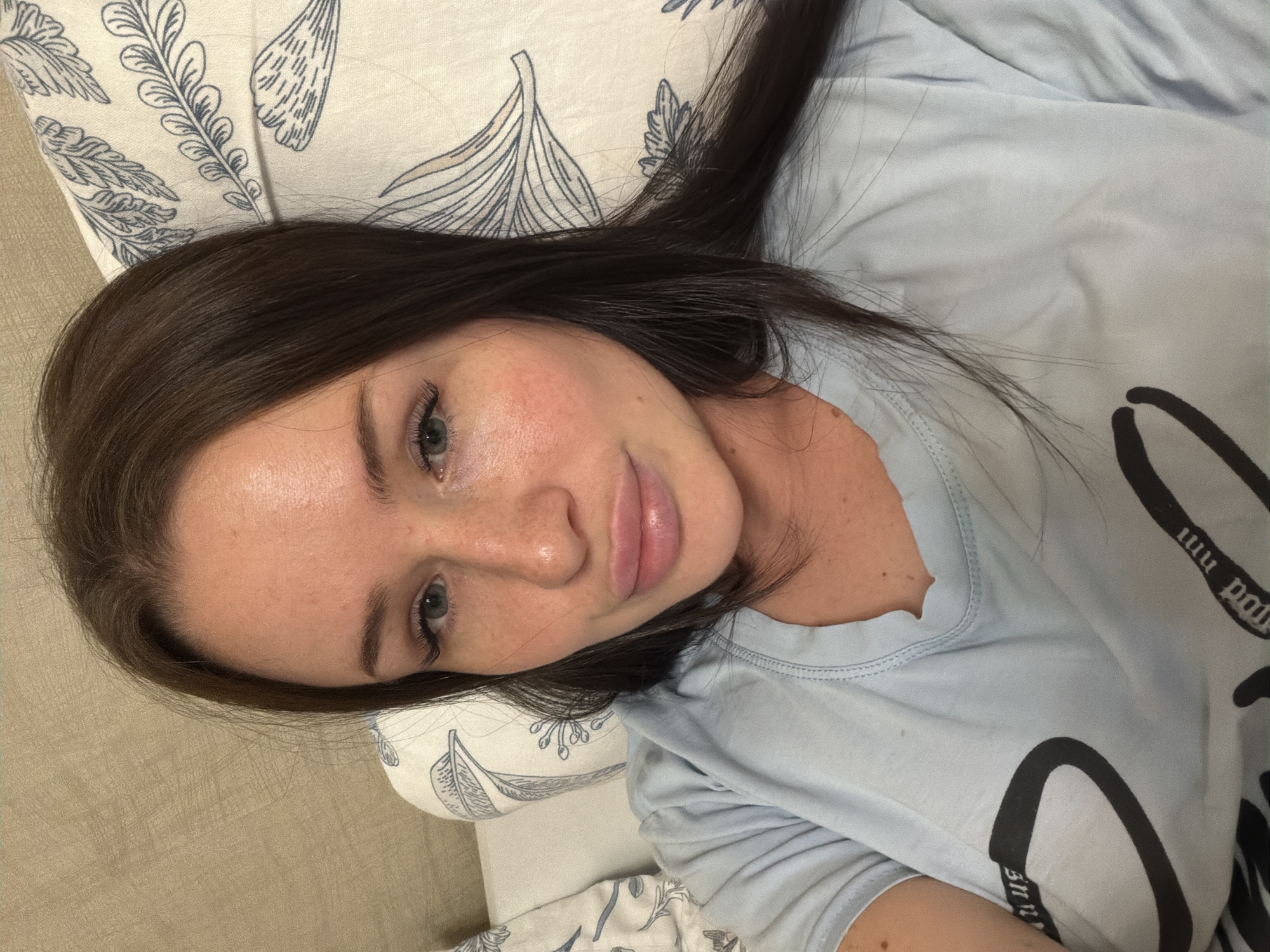 A young woman with long, dark brown hair is captured in a close-up, top-down perspective, lying on what appears to be a bed or couch. She is looking directly at the camera with a soft, calm expression, featuring a slight smile. Her hair is spread out behind and around her head, resting on a pillow and her shoulders. She wears a light blue t-shirt that features fragmented dark lettering or a graphic design; parts of letters like "nm" and "pou" are visible, along with a large curved shape.
Her skin has a natural appearance with some freckles visible on her nose and cheeks. Her eyes are open, and she has visible eye makeup, including eyeliner. Her lips are full, and she has well-defined eyebrows. She is resting her head on a patterned pillow, which is off-white or cream-colored with elegant blue or grey leaf and feather motifs. To the left of the pillow, a plain light brown or beige textured fabric, likely a bedspread or another part of the furniture, is visible. The scene is indoors, suggesting a relaxed setting, possibly a bedroom. No specific location cues for Saint Petersburg, Russia, are discernible from the image itself.