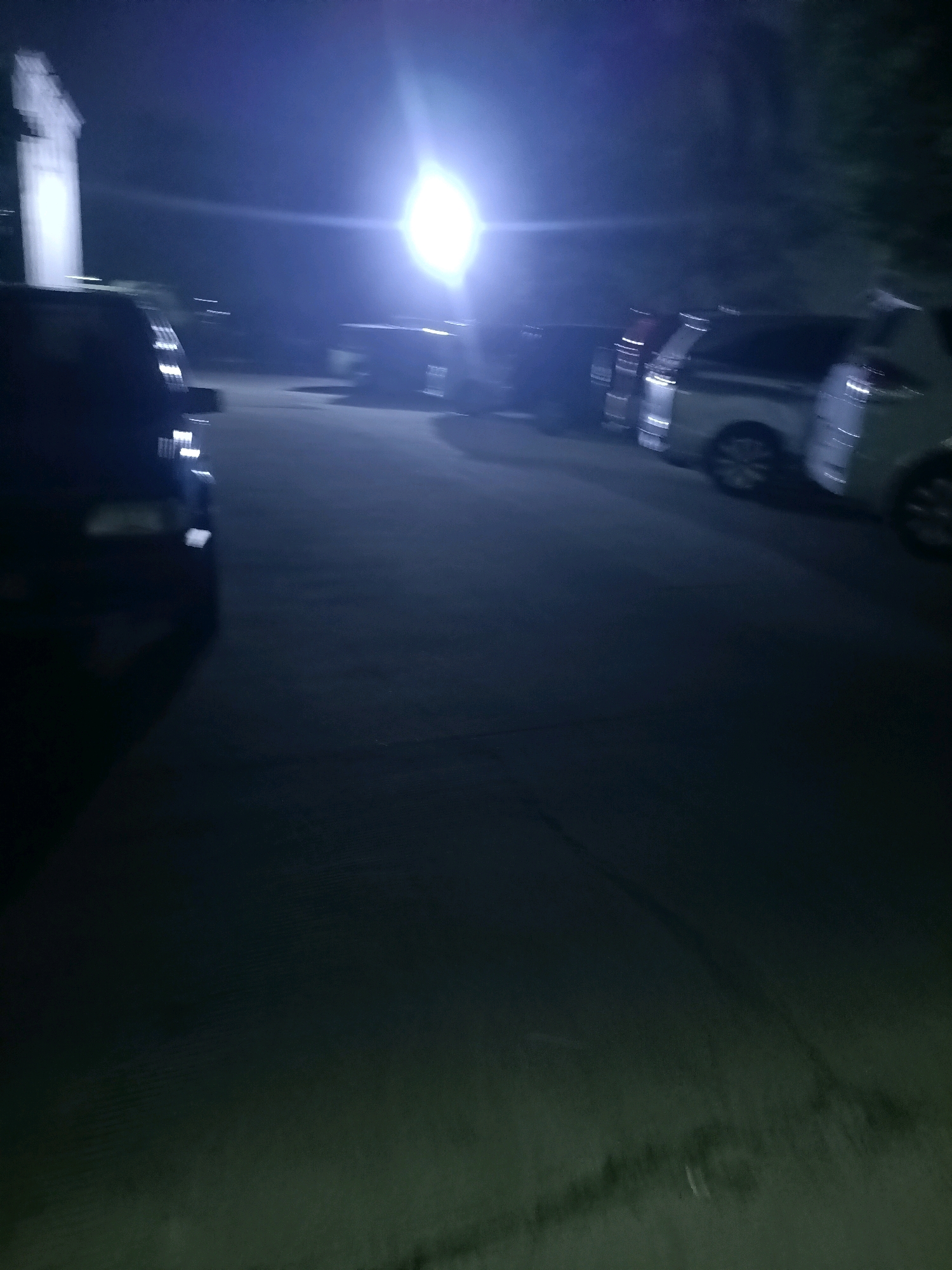 This image shows a dimly lit parking lot at night, likely in Karawang, Indonesia, with a bright light source in the background illuminating several parked vehicles. The lighting creates a hazy, blueish glow, obscuring fine details. In the foreground, a dark-colored vehicle is visible on the left. To its right, a line of other vehicles, possibly minivans or SUVs, are parked. The surfaces appear wet, reflecting the light. Trees or foliage are dimly visible in the background to the right. A tall, white structure, possibly a building or monument, is partially visible on the far left. The overall impression is a quiet, nocturnal scene in a semi-urban or commercial area.