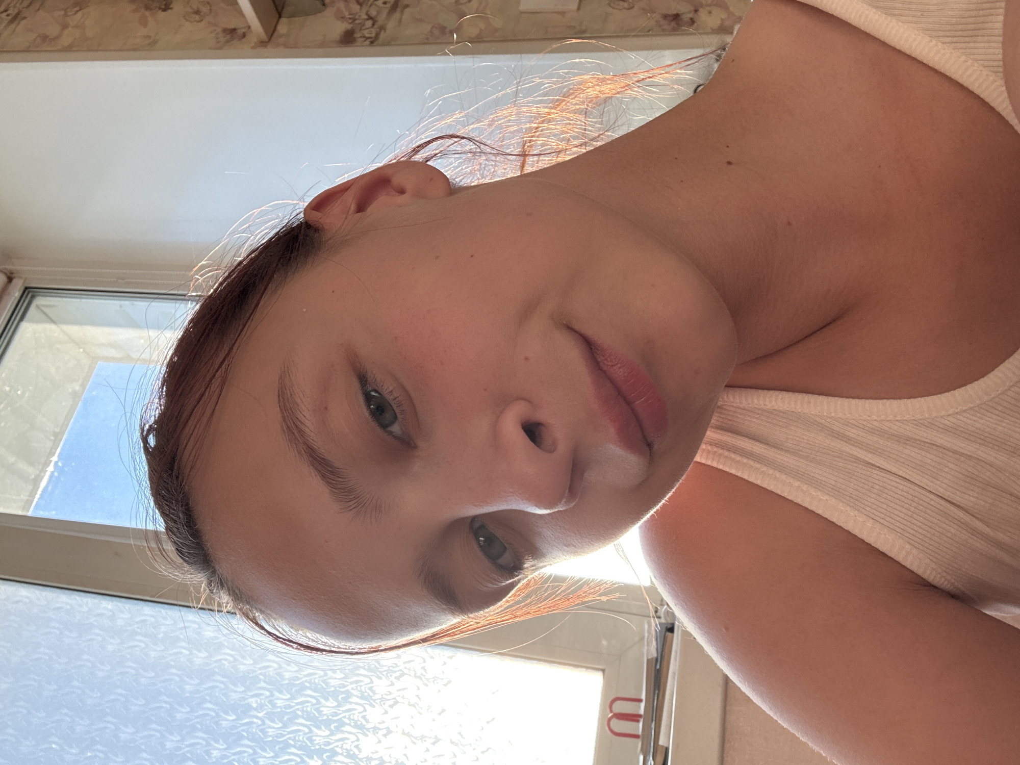 A young woman with reddish-brown hair is shown in a selfie taken indoors, with natural light coming from a window on the left. She is wearing a white ribbed tank top and looking slightly off-camera with a faint smile. Her hair is wispy around her face and catches the light. The background shows a window with a textured glass and a wall with a floral wallpaper pattern. On a surface near the window, there are some books and a small red object that resembles a U-shaped binder clip. The scene appears to be a casual, personal space, potentially a room in a residential building. The woman's skin tone is fair, and she has green eyes. The overall lighting suggests it is daytime, possibly morning or late afternoon, given the angle of the sunlight. There is no specific information about the location within Прикамский, Russia, as the image only provides visual cues.