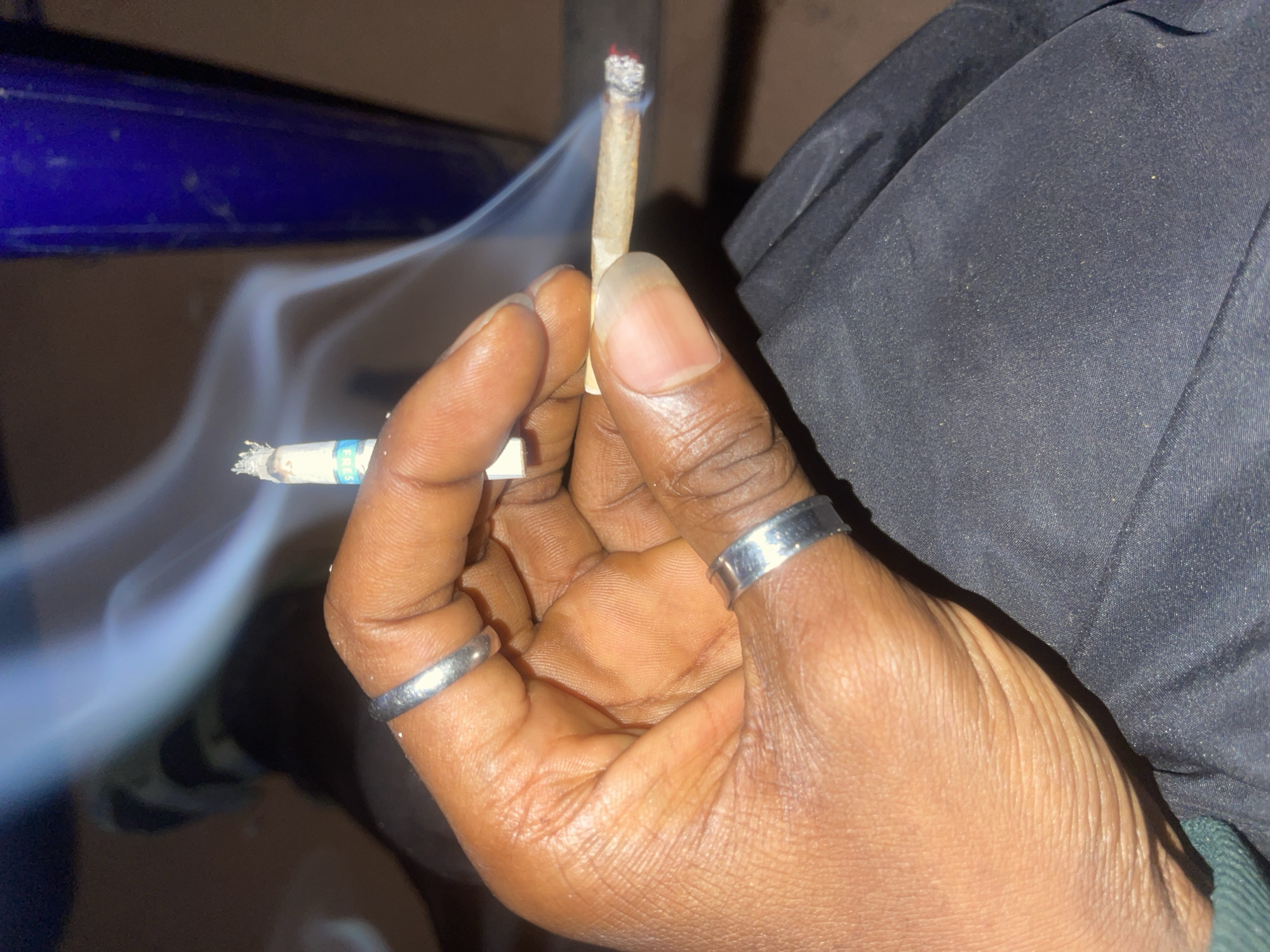 The image displays a close-up, top-down view of a dark-skinned human hand holding two lit smoking items, situated in Gura Top, Nigeria. The hand wears two plain, silver-colored rings: one on the index finger and another on the ring finger. Between the index and middle fingers, a partially consumed factory-made cigarette with a white filter and a blue band is held, actively producing a plume of white smoke. Simultaneously, the thumb and index finger hold a hand-rolled cigarette, also lit and emitting smoke, with a glowing red ember at its tip. The hand rests on dark, possibly black, fabric which appears to be clothing, with some visible lint or dust particles. The background is dark and out of focus, with a blurred blue vertical object visible on the right side, suggesting an indoor or dimly lit outdoor setting.