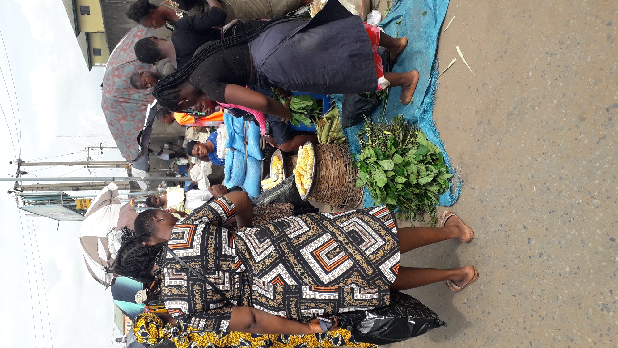 This is a medium shot, eye-level, outdoor photograph taken at a market in Onitsha, Nigeria. In the foreground, a woman wearing a black and gold patterned dress and beige sandals is standing facing away from the camera, holding a black plastic bag. To her right, a woven basket filled with corn on the cob is placed on a blue tarp. Next to the basket are bundles of green leafy vegetables. Behind the basket, another woman in a dark top and dark skirt is interacting with the vegetables, also facing away from the camera. Several other people are visible in the background, including a woman in a blue shirt sitting behind a table laden with blue sacks, and another woman in a yellow dress standing near a stall with umbrellas. The environment appears to be a bustling marketplace with various stalls, tarps, and overhead structures, including utility poles and a billboard. The ground is paved with concrete.