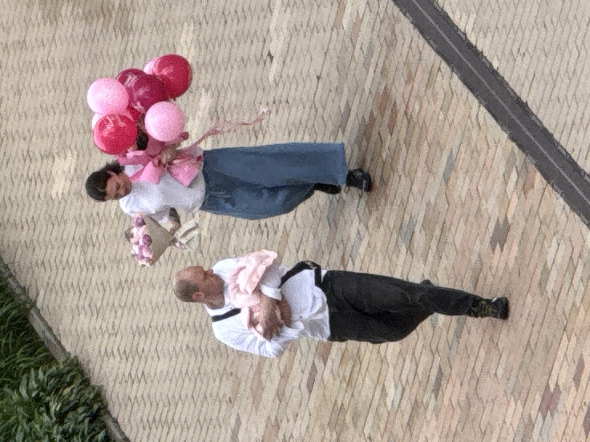 In Khimki, Russia, a man and a woman walk on a brick-paved pathway. The man, bald and wearing a white long-sleeved shirt and dark pants, carries a baby wrapped in a pink blanket. He has a black strap across his chest. The woman, with dark hair pulled back, wears a white top and wide-leg blue pants. She holds a bouquet of pink and purple flowers in one hand and a bunch of pink balloons in the other. Green foliage is visible along the upper left edge of the image.