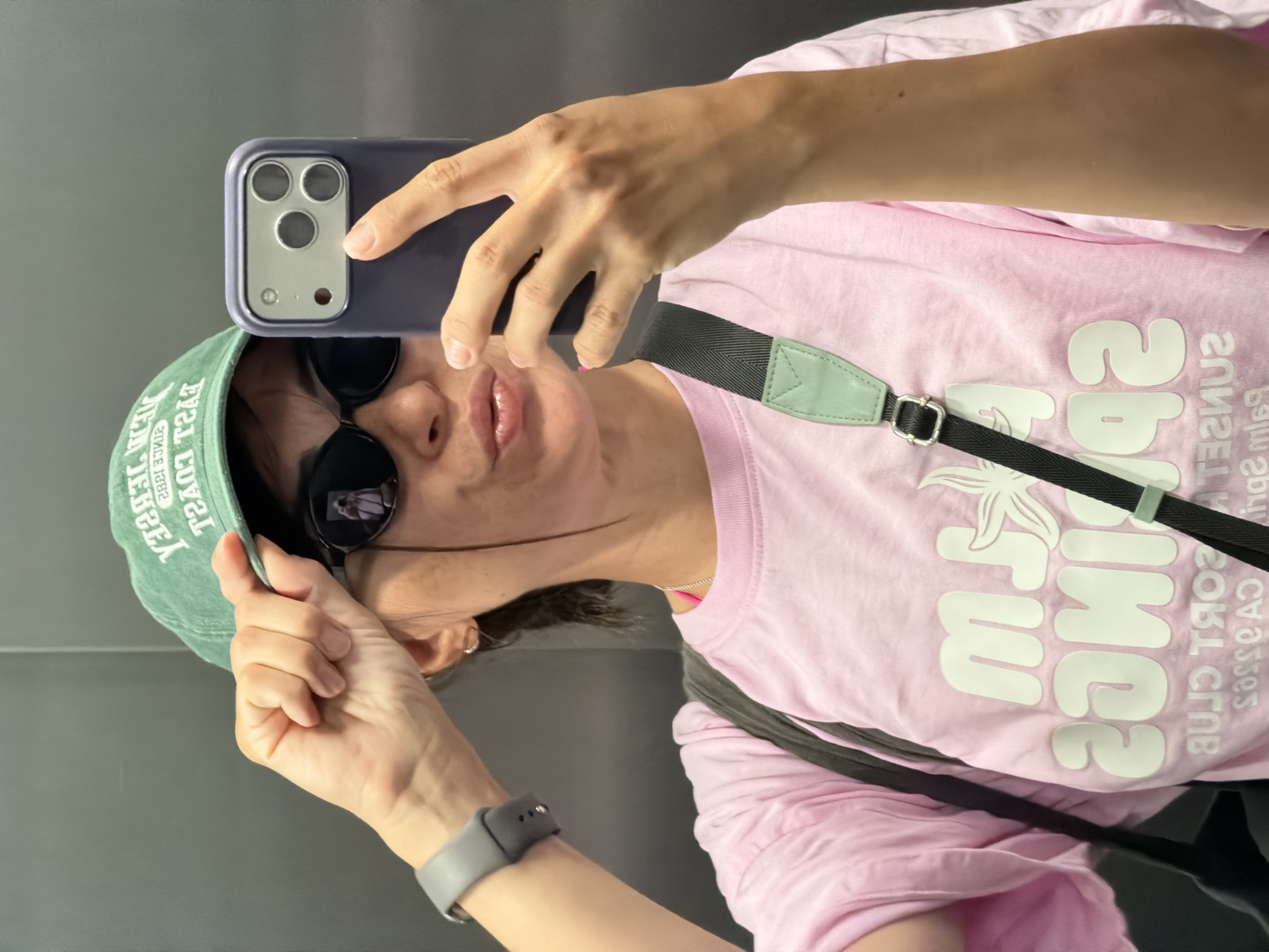 A woman is taking a selfie with her phone in an elevator with a gray wall. She is wearing a pink t-shirt, sunglasses, a green baseball cap, and a gray Apple Watch. The writing on her t-shirt says "Sunset Resort Club Budva Montenegro." The hat says "New Jersey Coast 1982" in white.