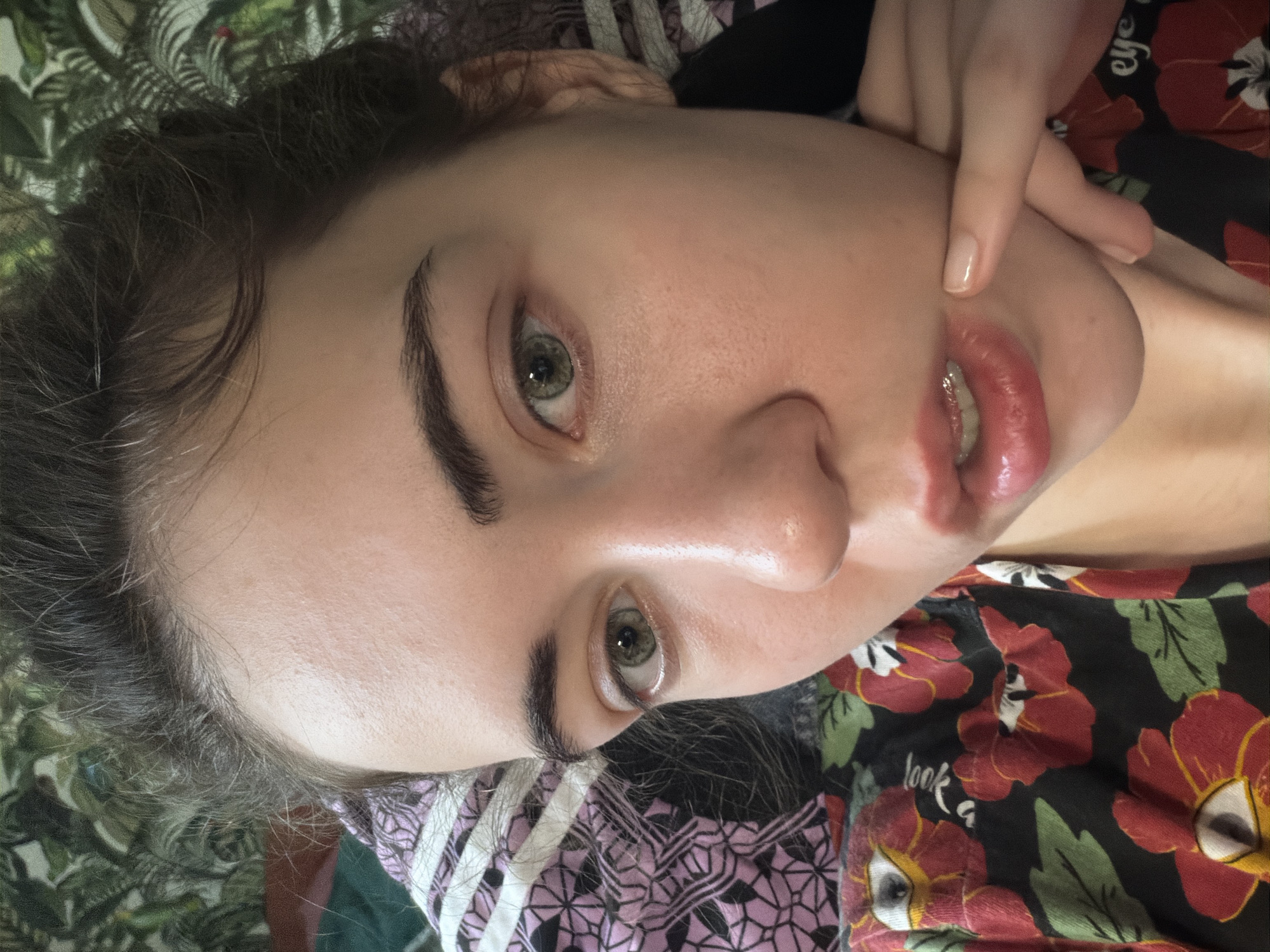 A young woman is lying down, her face close to the camera. Her hair is dark and messy, partially obscuring her forehead and the top of her head. She has light skin, green eyes, and dark eyebrows. Her lips are slightly parted, revealing her teeth, and they appear to be glossed. A finger is gently touching the side of her face near her lips. She is wearing a black shirt with a pattern of red flowers and green leaves, and a pink and white striped fabric is visible behind her head. The background appears to be bedding with a floral pattern. The scene is indoors. There is no information about the location in Zavodoukovsk, Russia.