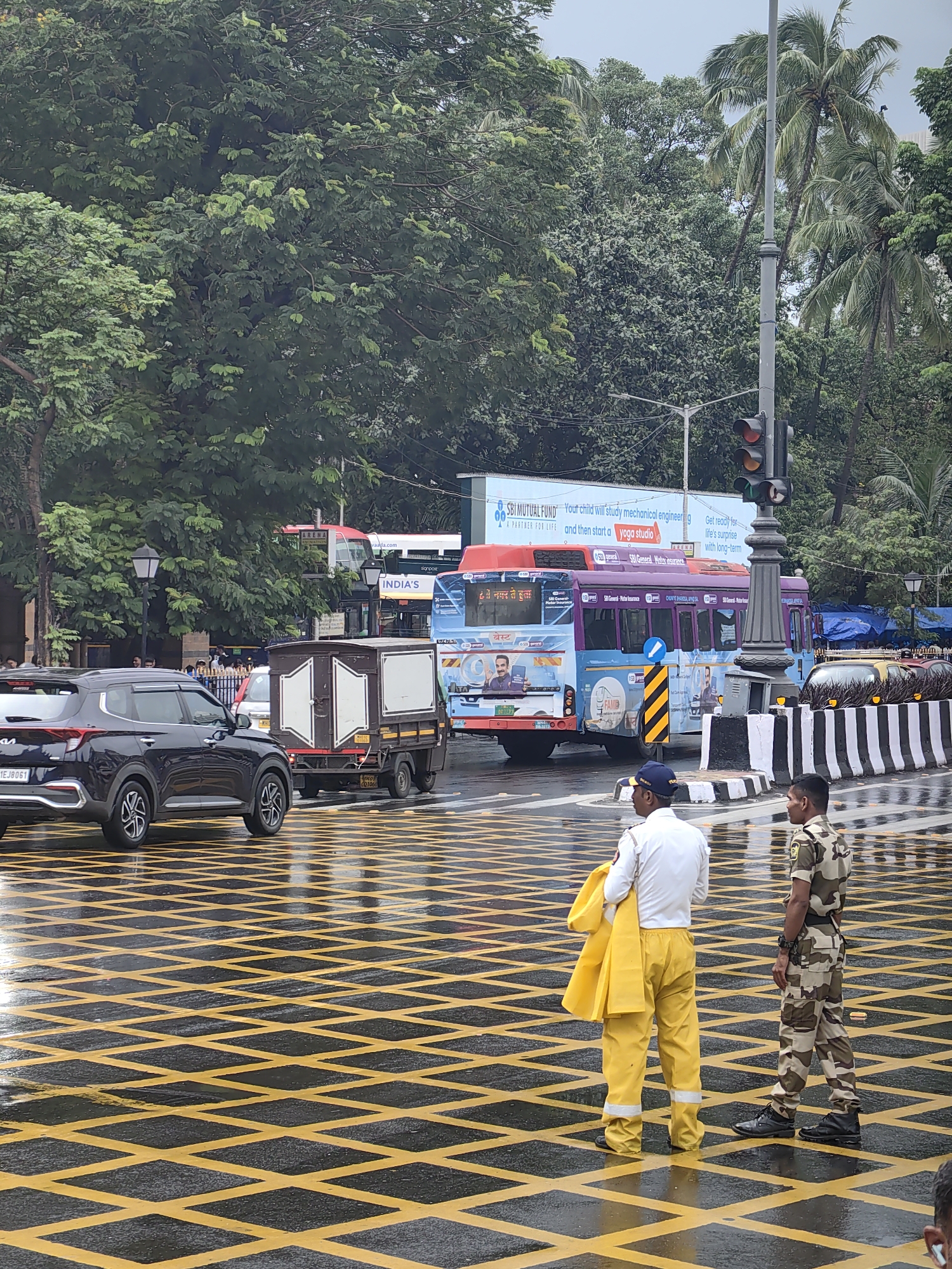 The image depicts a street scene in Mumbai, India, likely during or after rainfall, evidenced by the wet road surface marked with yellow criss-cross patterns. Two men in uniform are visible in the foreground, standing on the marked road. One man, on the left, wears a white shirt, yellow pants, and a blue cap, and holds a yellow garment over his shoulder. The other man, on the right, is dressed in a camouflage uniform. In the background, several vehicles are present, including a black Kia Carens car, a small truck with a brown box-like cargo area, and two buses. One of the buses is predominantly blue with purple accents and features advertisements, including one for "SBI Mutual Fund." A traffic light is visible on a pole, with the red and yellow lights illuminated. Lush green trees and palm trees line the background, suggesting an urban environment with significant vegetation. A large billboard advertising "SBI MUTUAL FUND" is also prominent. The overall atmosphere suggests a busy intersection in a tropical climate.