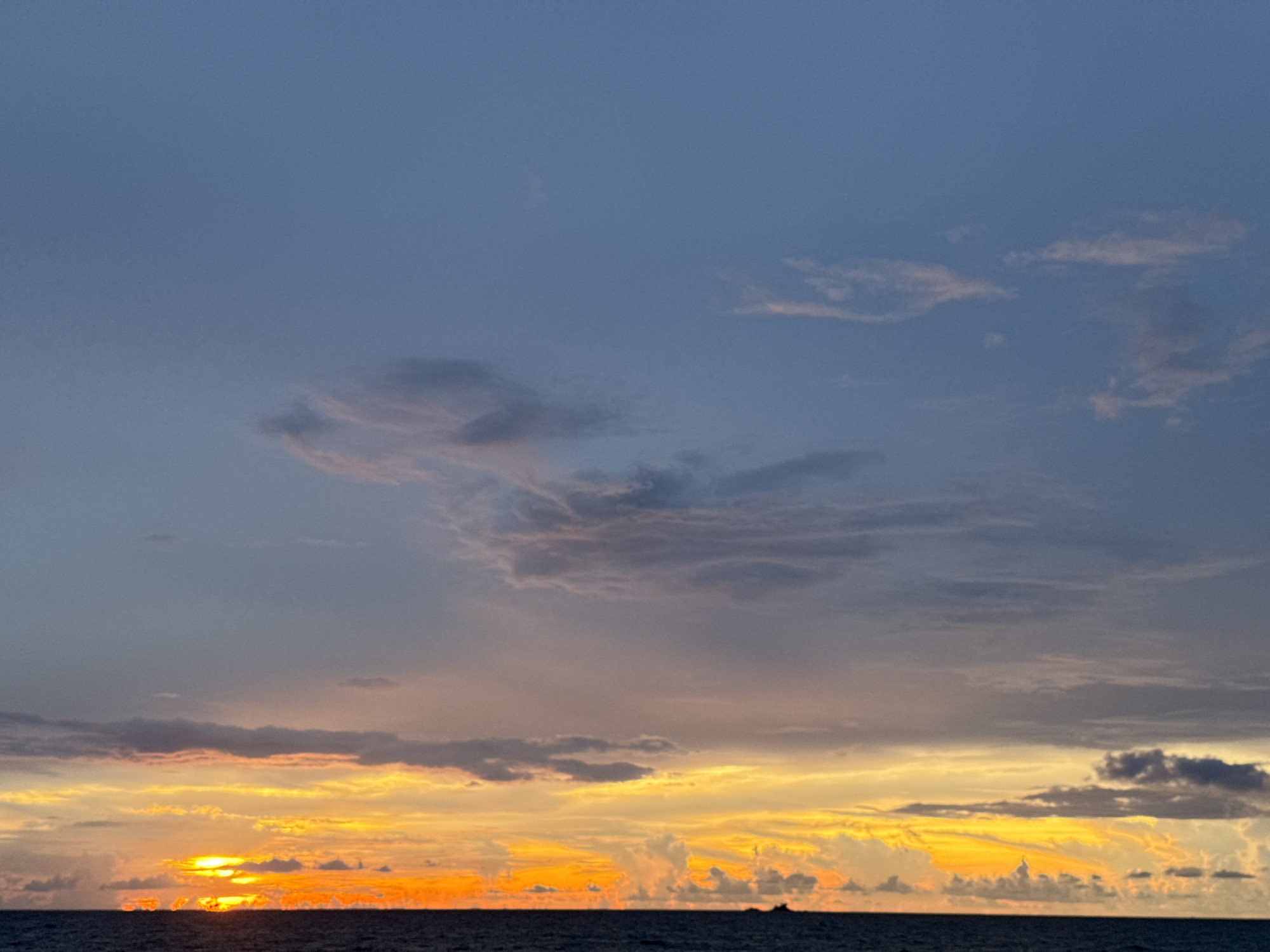 The image depicts a sunset over the sea. The sky is a gradient of colors, with shades of blue, purple, orange, and yellow. The sun is low on the horizon, casting a warm glow across the scene. Clouds are scattered across the sky, adding texture and depth to the image. The sea is dark and calm, with a thin line of land visible in the distance. The image was taken in Choeng Thale, Thailand, a location known for its beautiful beaches and sunsets.