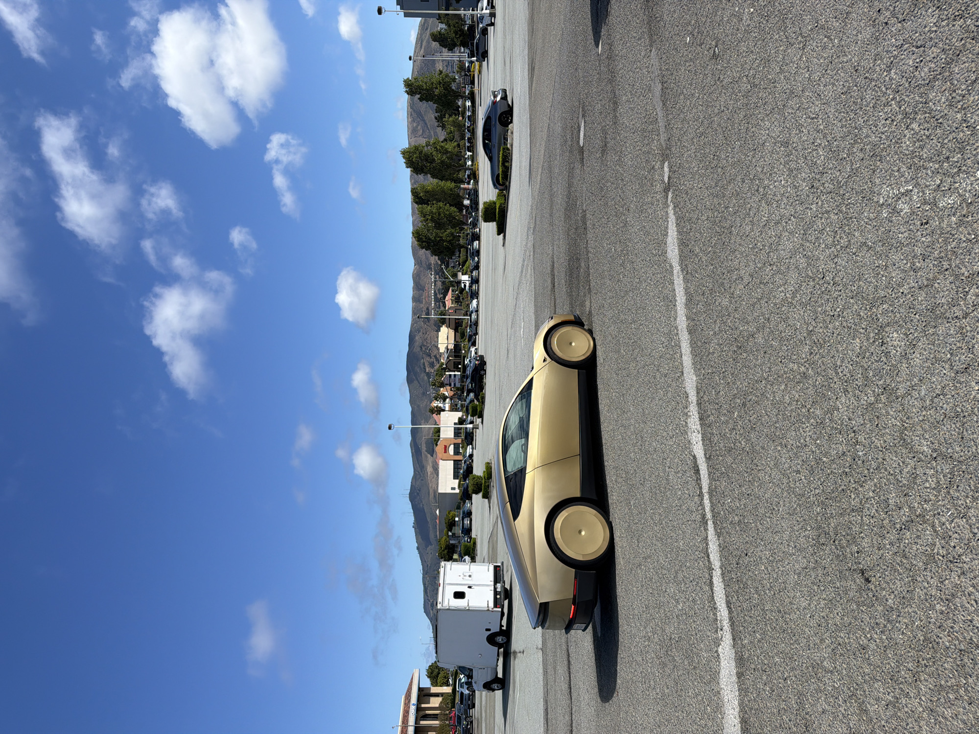 This media file captures an outdoor scene in a parking lot, likely in San Bruno, USA, given the context. The primary focus is a futuristic-looking gold car with unique, disc-like wheel covers, parked in the foreground. Behind the gold car, several rows of other vehicles are visible, suggesting a dealership or a large parking area. In the middle ground, there are trees and buildings, possibly part of a commercial area or shopping center. A white box truck is also present. The background features rolling hills or mountains under a clear blue sky dotted with fluffy white clouds. The lighting suggests it is daytime, likely a sunny afternoon. There is no visible text in this media file. The overall impression is that of a bright, possibly warm day in a suburban or commercial setting with an eye-catching vehicle.