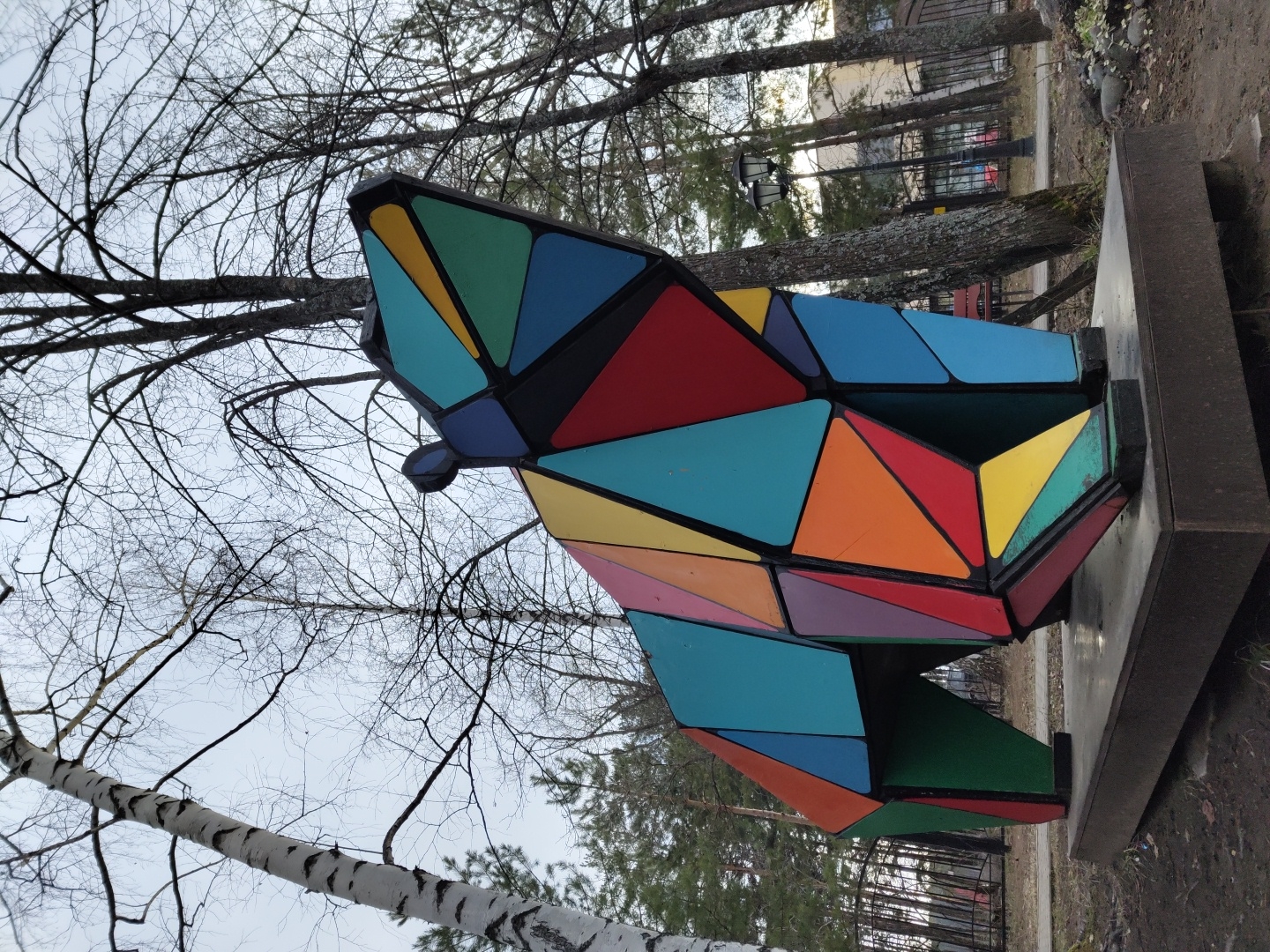 The image captures a vibrant, multi-colored geometric sculpture of an animal, possibly a bear or wolf, situated outdoors in what appears to be a park or public space in Chaikovsky, Russia. The sculpture is constructed from numerous triangular and quadrilateral panels, rendered in a bright palette of red, blue, green, yellow, orange, turquoise, and purple, all outlined by dark, possibly black, frames. It stands on a dark, rectangular base.
The scene is set on an overcast day, evident from the soft, diffused lighting and the pale, uniform sky. Many deciduous trees in the background and overhead are bare, their branches forming intricate patterns against the sky, suggesting late winter or early spring. A distinctive birch tree with white, peeling bark is visible on the left side. Further back, evergreen trees, lampposts, a path, and distant buildings indicate an urban or semi-urban environment. No specific activity or event is occurring; the image focuses solely on the static sculpture within its natural surroundings. There is no visible text in the media file. The overall impression is one of calm and artistic presence within a natural, albeit human-influenced, landscape.