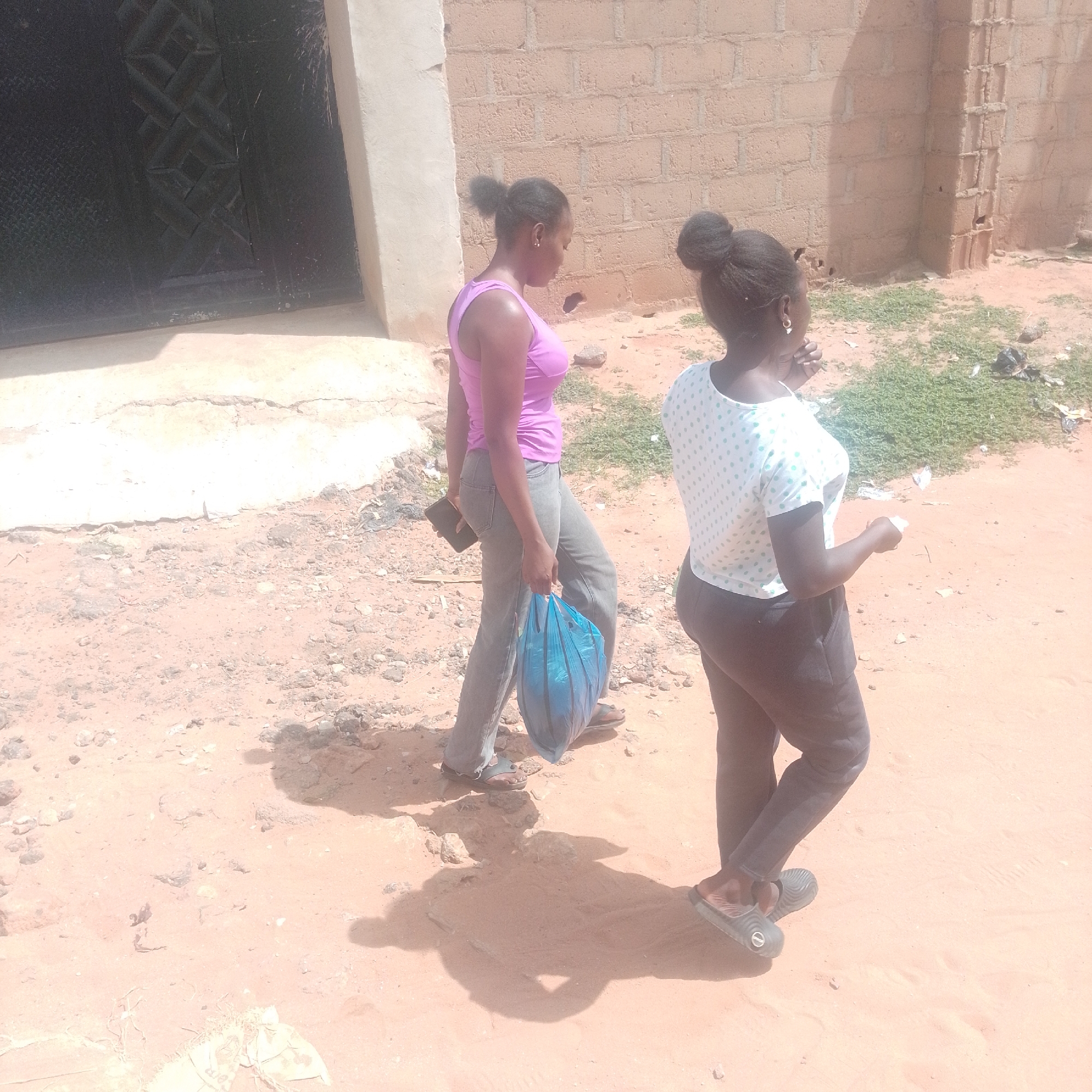 Two women are walking in an outdoor environment. The woman in the foreground on the right is wearing a white t-shirt with light blue polka dots and dark pants. She has her hair pulled back in a bun. The woman in the background on the left is wearing a pink tank top and grey pants. She is carrying a blue plastic bag in her right hand and a dark object, possibly a phone, in her left hand. Her hair is also pulled back in a bun. They are walking on a sandy, dirt path next to a brick wall. There is some sparse green vegetation on the ground. The area appears to be an urban or semi-urban location with a building to the left, featuring a dark, patterned door.