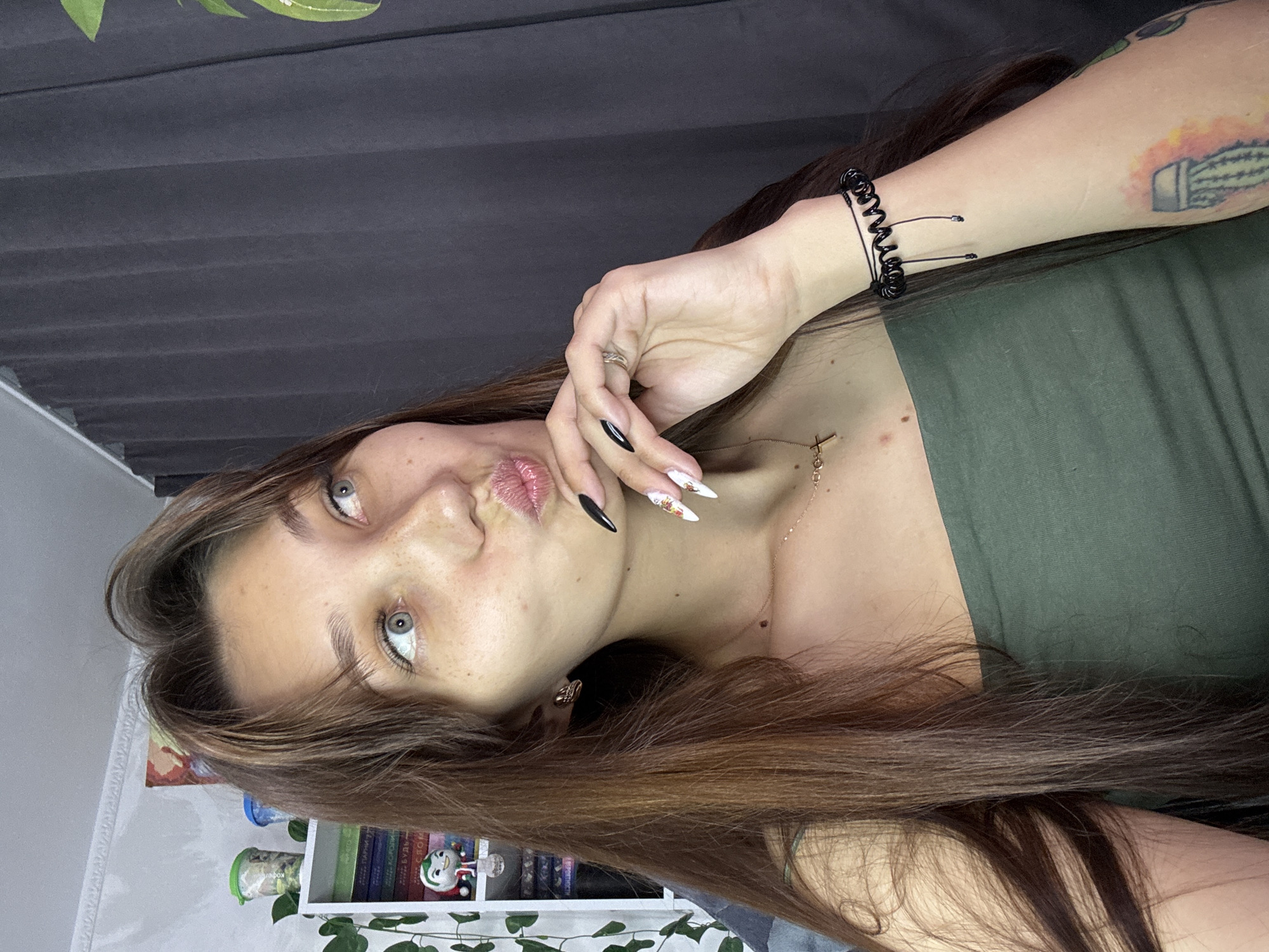 A young woman with long brown hair and fair skin, adorned with freckles, is captured in a medium shot, posing with her hand on her chin and lips puckered. She is wearing a dark green strapless top and a delicate gold necklace with a cross pendant. On her left wrist, she sports a black coiled bracelet. The background features a dark gray curtain and a bookshelf filled with books, a plant, and decorative items, suggesting an indoor setting, possibly a bedroom or study. There is no information in the image that allows for the determination of its location within the city of Novokuznetsk, Russia.