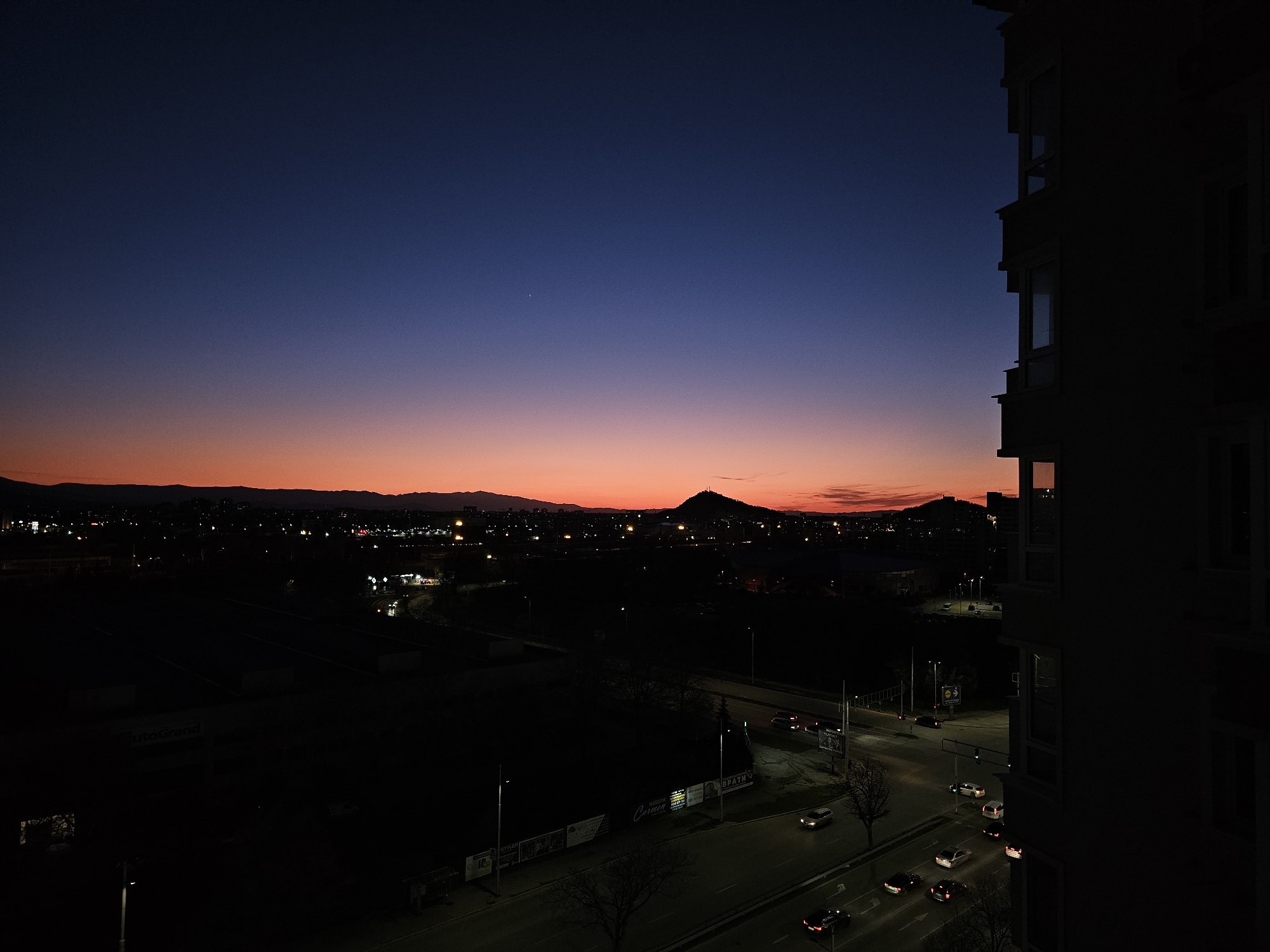 The image depicts a nighttime cityscape in Plovdiv, Bulgaria, at twilight. A multi-story building is visible on the right side of the frame, its dark silhouette contrasting with the illuminated windows on its facade. A highway stretches across the lower part of the frame, with cars moving along the road, their headlights illuminating the asphalt. Streetlights are also present, casting a glow on the scene. In the background, a silhouette of a mountain range is visible against the colorful sky, which transitions from dark blue to orange and red hues. The city lights of Plovdiv are scattered throughout the landscape.