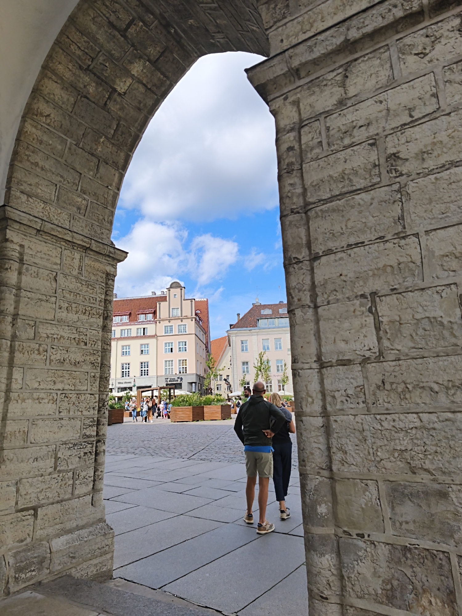 This is a low-angle, full shot of an archway looking out onto a town square in Tallinn, Estonia. The archway is made of light-colored stone blocks. Through the archway, a plaza with cobblestone paving is visible, with several people walking and standing. In the background, there are buildings with red tile roofs and light-colored facades, characteristic of Old Town Tallinn. A couple, a man and a woman, are walking away from the camera, through the archway. The man is wearing a dark jacket and light shorts, and the woman is wearing dark pants and a dark shirt. The sky is blue with some white clouds. Wooden planters filled with greenery are placed around the plaza.