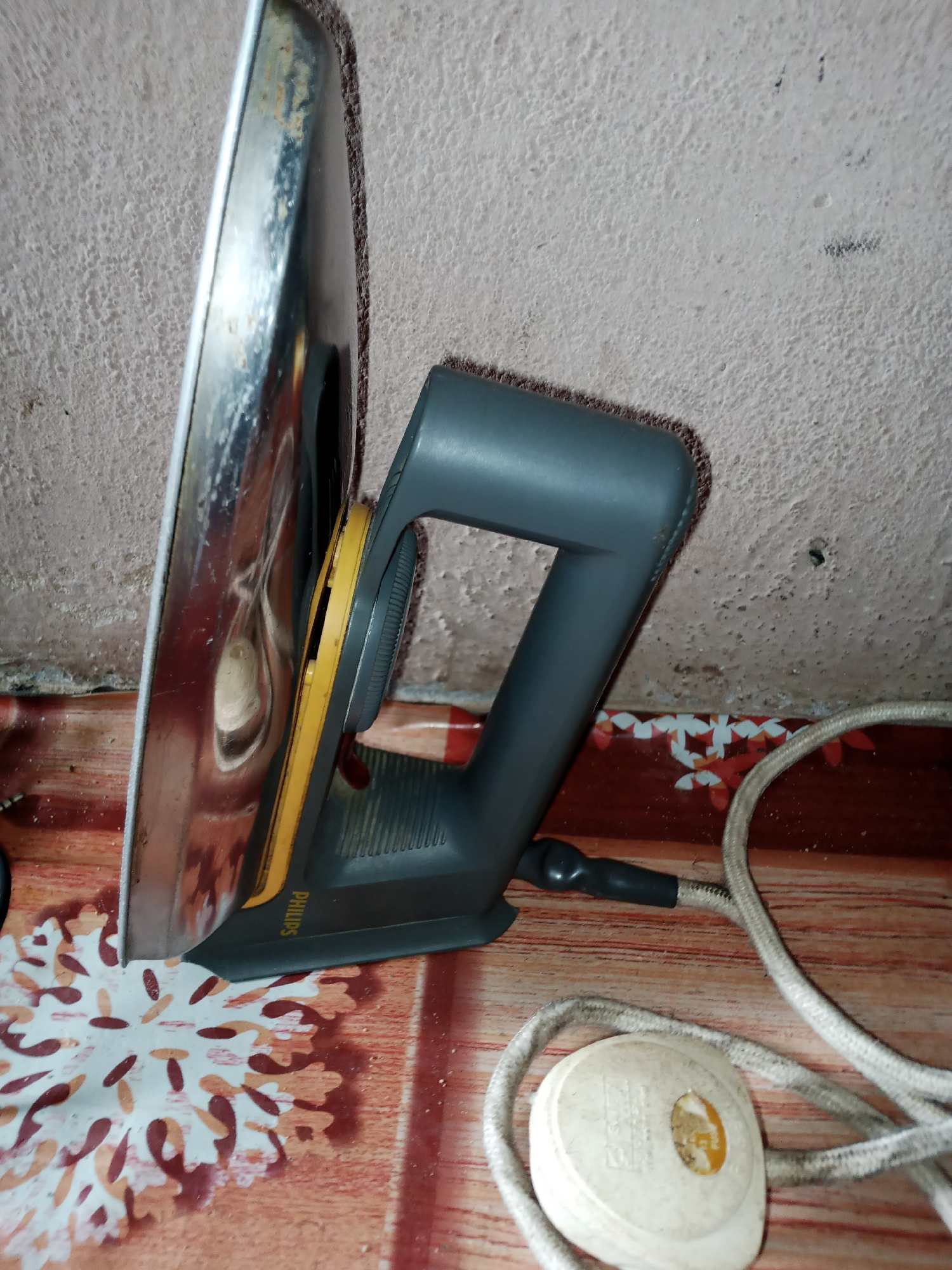 A close-up shot shows a vintage Philips iron. The iron is primarily gray with a yellow dial. It rests on a surface with a reddish-brown wooden grain and a decorative pattern of brown and tan floral motifs. To the right of the iron, a white power cord is visible, coiled and leading to a white electrical plug. The background is a textured, light-colored wall, possibly concrete, with some darker marks and imperfections. The scene appears to be indoors, with the lighting suggesting it could be daytime. The overall impression is one of a functional, yet well-used household item in a simple setting. The text "PHILIPS" is visible on the yellow part of the iron.