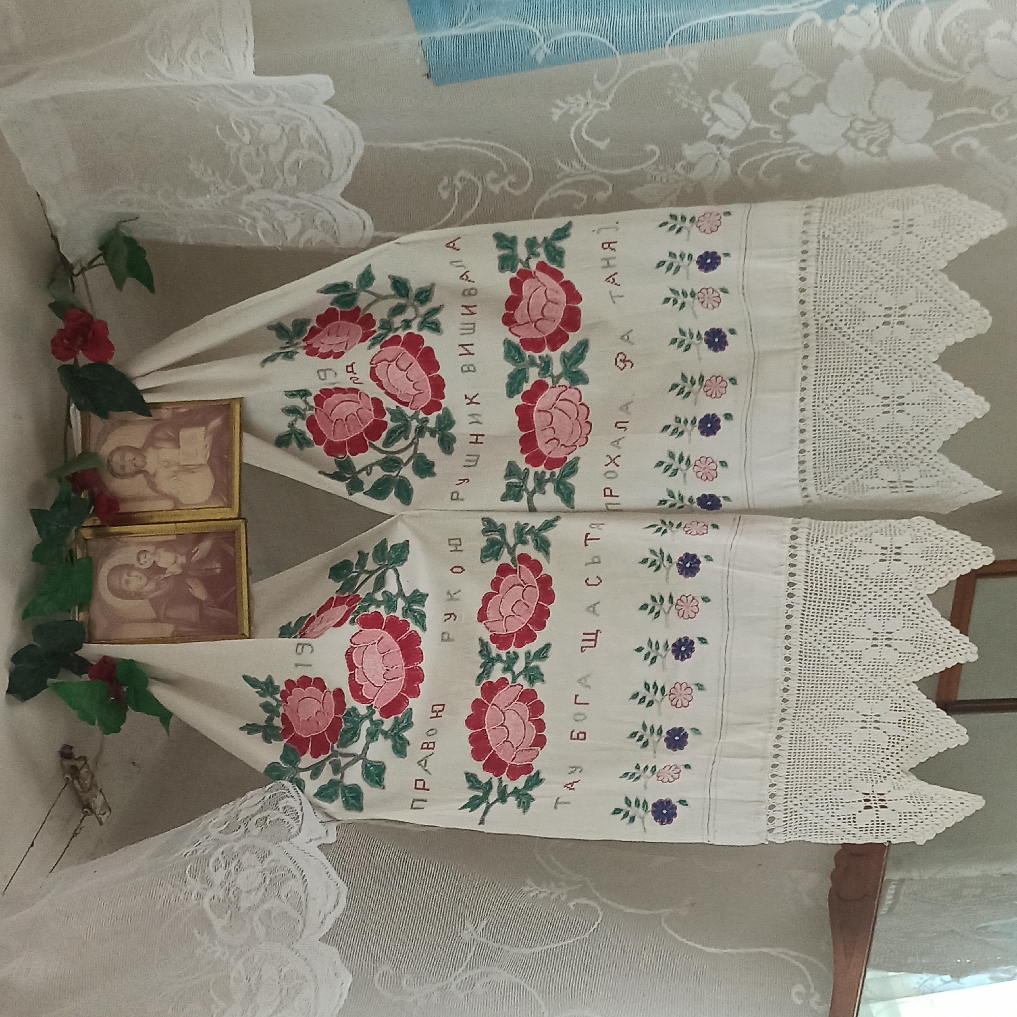 This is a close-up, indoor, eye-level shot of an embroidered towel hanging on a wall. The towel is white with floral embroidery in pink and green. There are two small religious icons hanging above the towel. The towel is decorated with Cyrillic writing. The background shows white lace curtains. The scene is likely a home or a religious establishment.