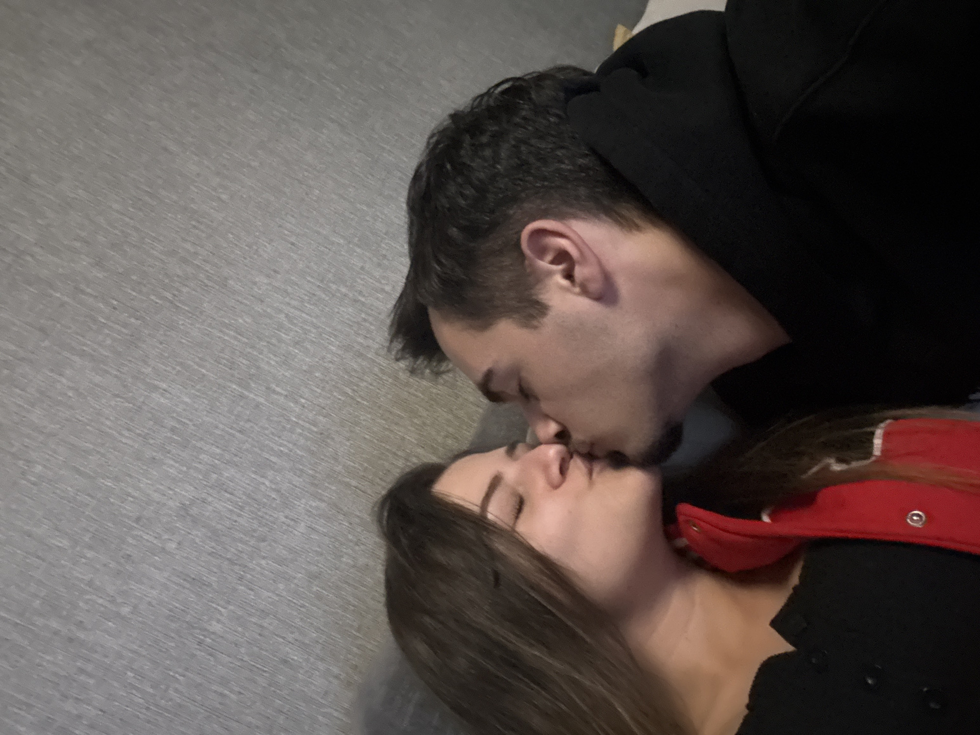 A man and a woman are lying on a gray surface and kissing. The man is wearing a black hoodie. The woman is wearing a black top with a red trim. The location is unknown, but it is not visibly identifiable as being in Sofia, Bulgaria.
