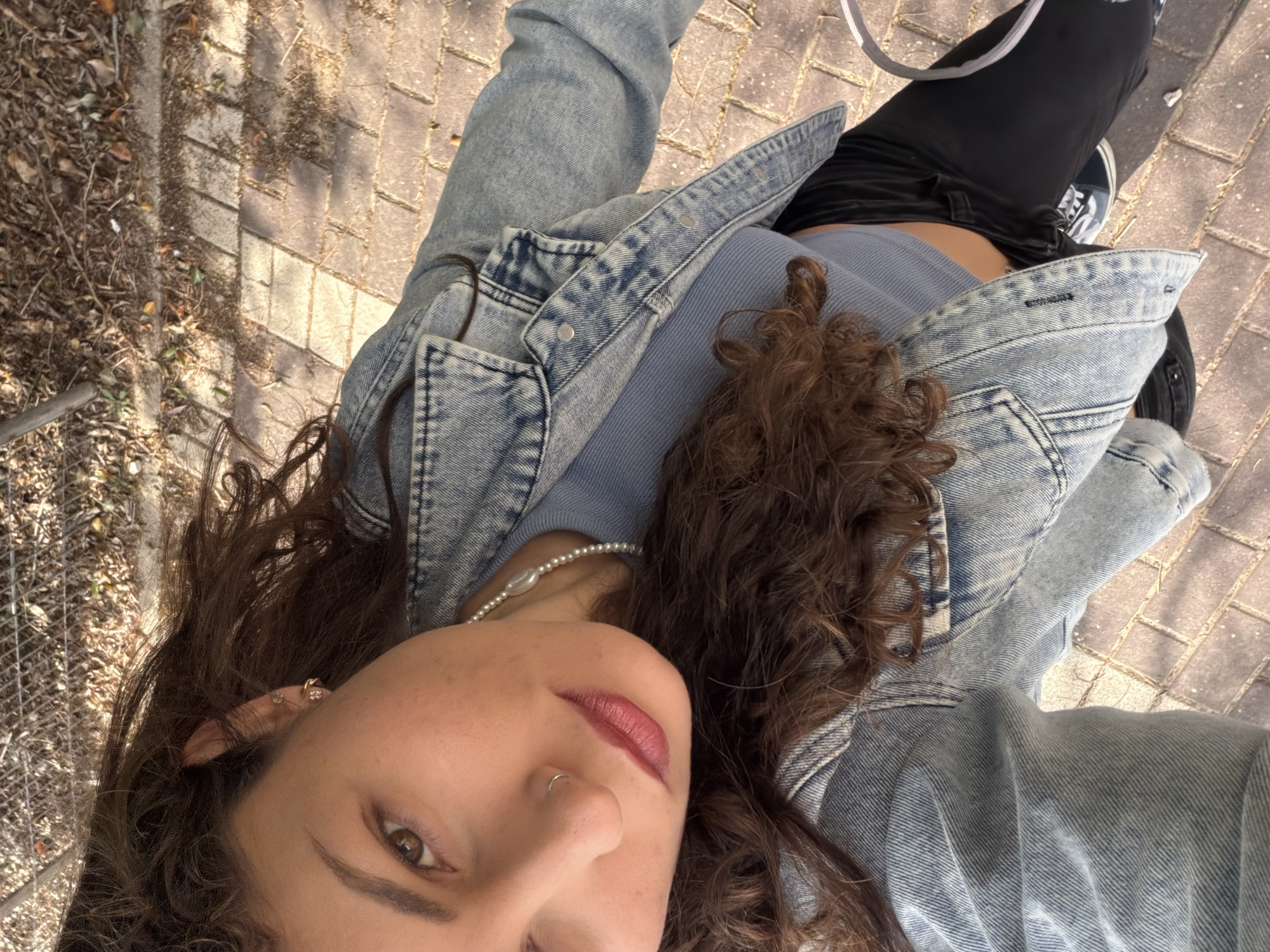 A woman with curly brown hair is pictured in a selfie taken outdoors. She is wearing a light blue denim jacket over a light blue ribbed top. She has a pearl necklace and a nose ring. The background includes a brick-paved area and some foliage. The location is potentially in Kiryat Bialik, Israel.