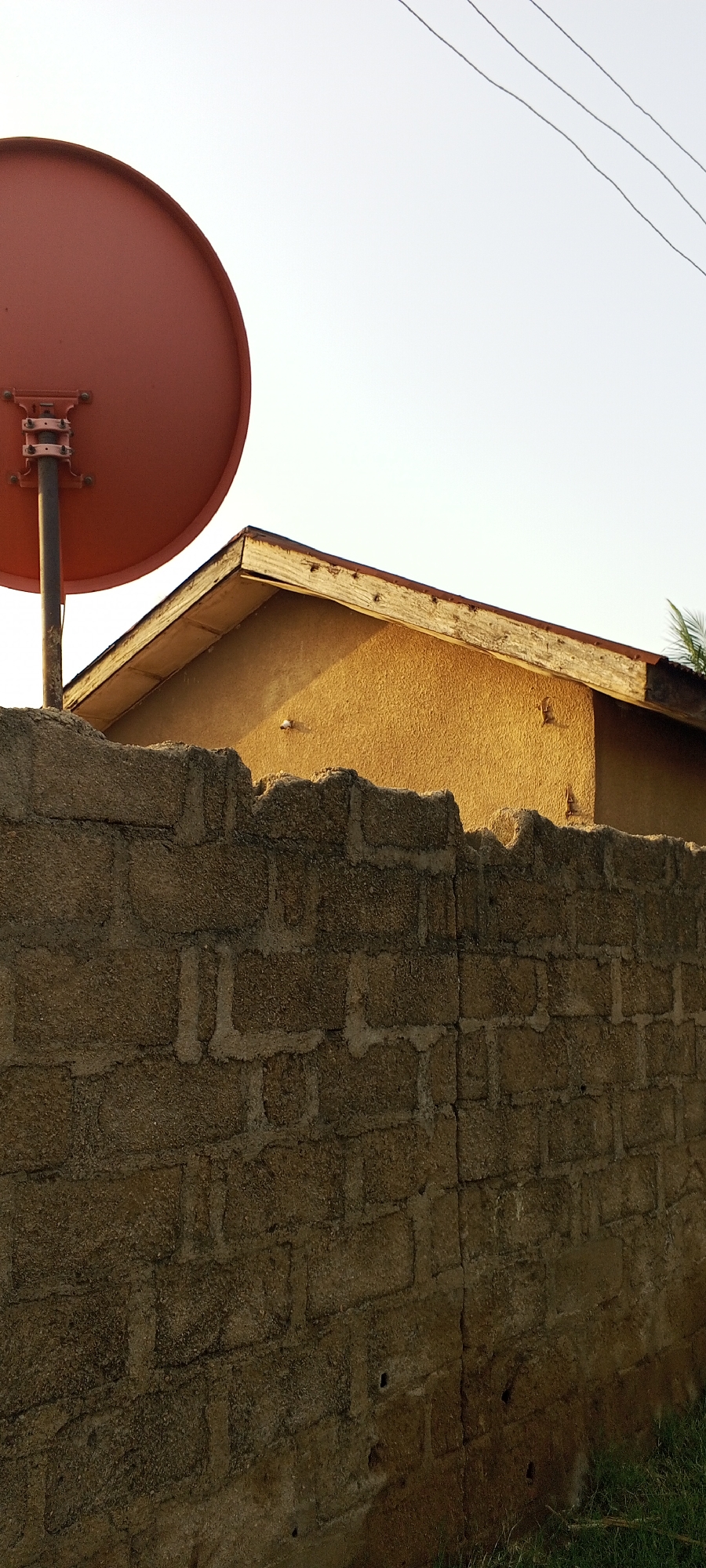 This image depicts an outdoor scene, likely in Jalingo, Nigeria, featuring a red satellite dish mounted on a pole, positioned above a textured, tan-colored building with a peaked roof. In the foreground, a roughly constructed wall made of large, interlocking blocks with visible mortar separates the viewer from the building and dish. The sky above is a clear, pale white, with three thin, dark power lines traversing the upper right quadrant. The sunlight casts shadows, indicating it is likely late afternoon or early morning. There are no people or animals visible in the image. The environment appears to be an urban or semi-urban setting with a focus on residential structures and utilities.