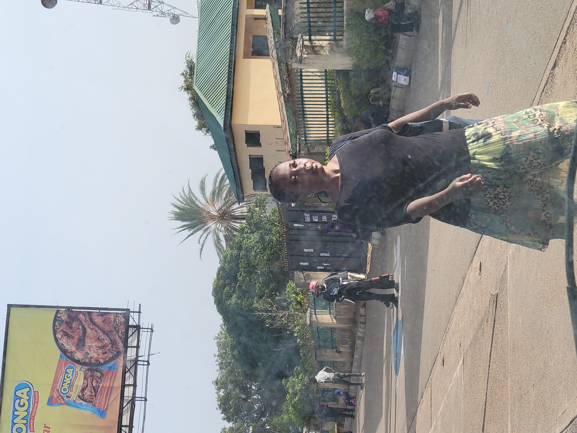 A woman is standing in the foreground on a street in Jos, Nigeria. She is wearing a black top and a green and brown patterned skirt. Behind her are trees, a building, and a billboard advertising ONGA chicken flavor. People are walking on the sidewalk in the background. A container is also visible behind the woman. A palm tree can be seen to the left of the building. A tall communication tower is visible in the upper left corner of the frame.