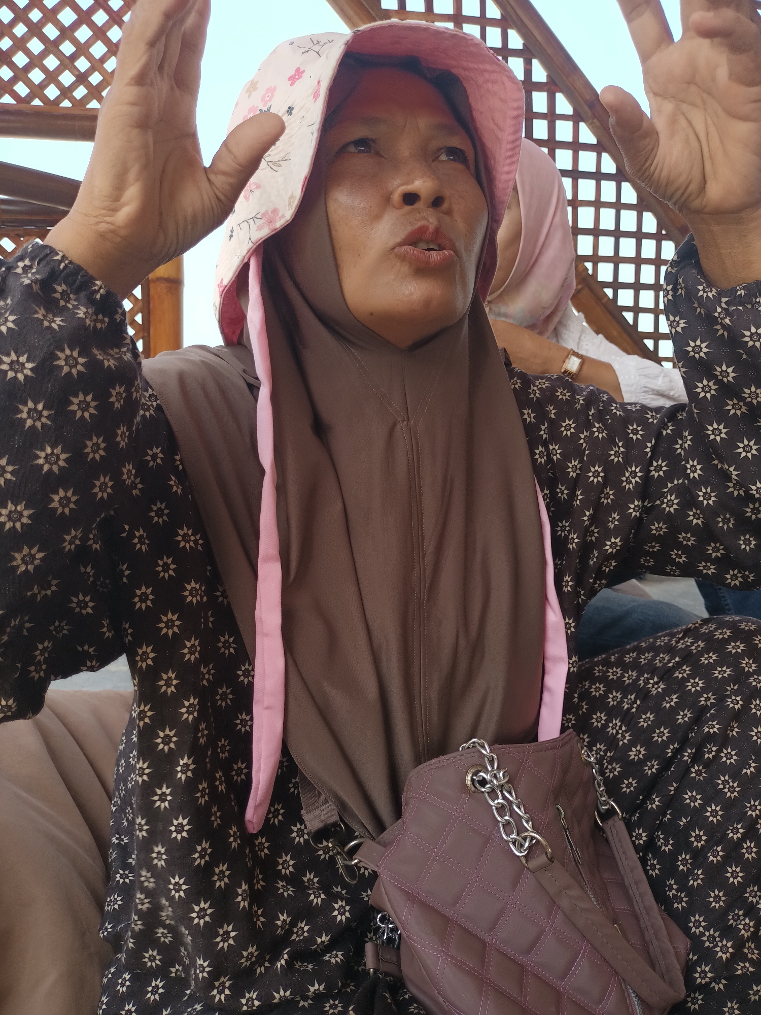 In Bulok, Indonesia, a close-up shot shows a middle-aged woman in the foreground wearing a brown hijab and a bucket hat adorned with pink flowers. She is dressed in a dark brown top with a white star pattern. Her hands are raised near her head, with one hand partially obscuring her face as she looks upwards and to the right, her mouth slightly open as if speaking. A quilted, dark pink purse with a silver chain strap hangs from her shoulder. In the background, another person wearing a light pink hijab and a white top is partially visible. The scene appears to be under a wooden lattice structure, suggesting an outdoor or semi-outdoor setting. The overall lighting is bright, indicating daytime.