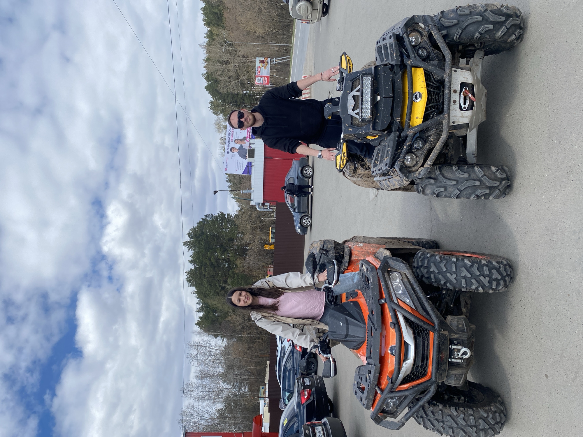 A man and a woman are sitting on two ATVs in the city of Chaikovsky, Russia. The man, wearing sunglasses and a black hoodie, sits on a yellow and black ATV. The woman, wearing a pink shirt and beige jacket, sits on an orange ATV. Both ATVs appear to be dirty, as though they have been driven off-road. Behind them, cars are parked and a large billboard is visible. The environment suggests they are in a parking lot or similar paved area, with trees and buildings in the background. The sky is cloudy.