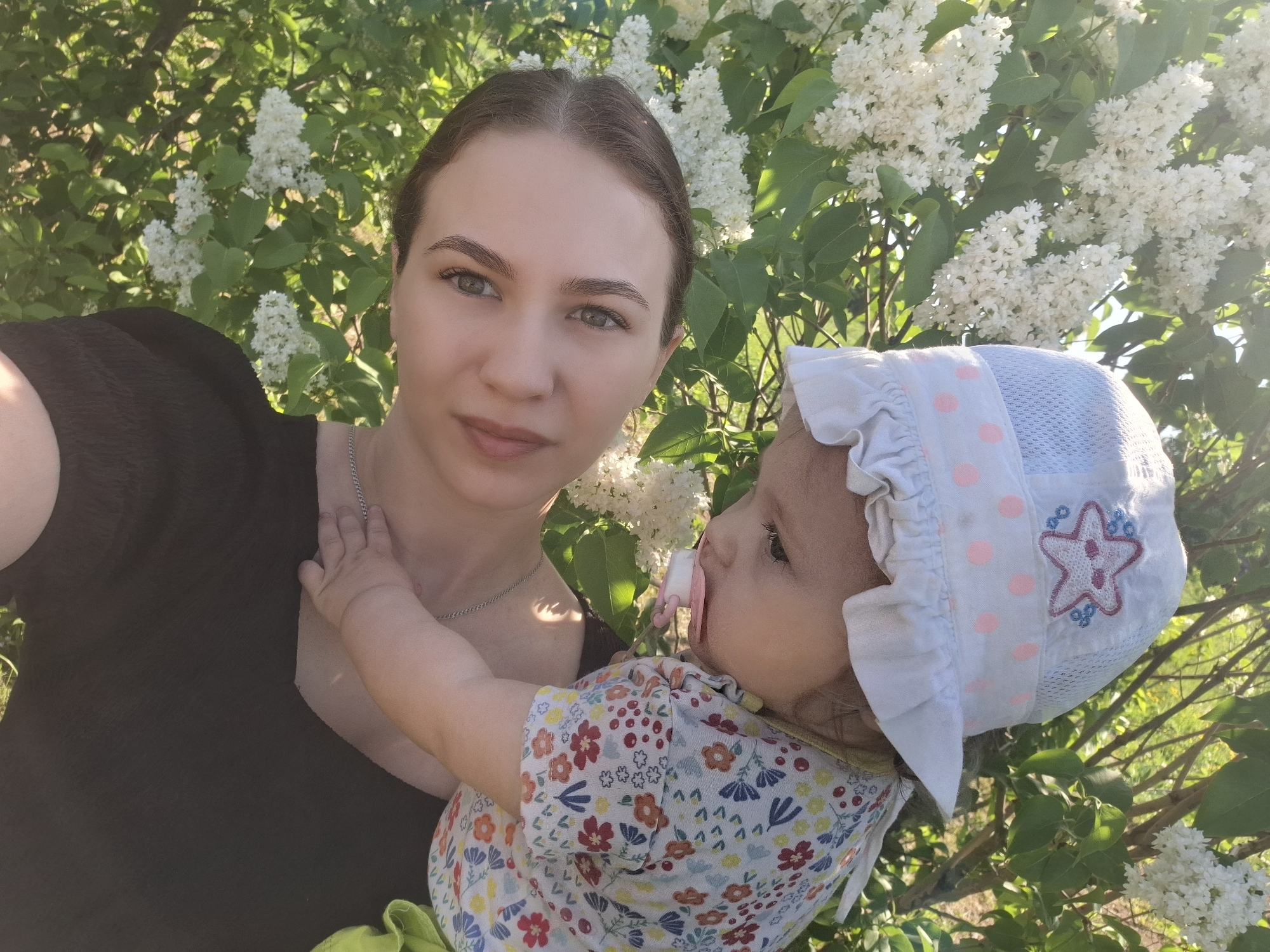 A woman is holding a baby in an outdoor setting. The woman, with her hair pulled back, is wearing a dark-colored top and a thin chain around her neck. The baby is wearing a patterned shirt, green bottoms, and a light-colored bonnet adorned with pink polka dots and a starfish design. The baby has a pacifier in its mouth and one hand resting on the woman's chest. White flowering bushes surround the pair, and sunlight filters through the foliage. The location is Nizhnyaya Gar', Russia.