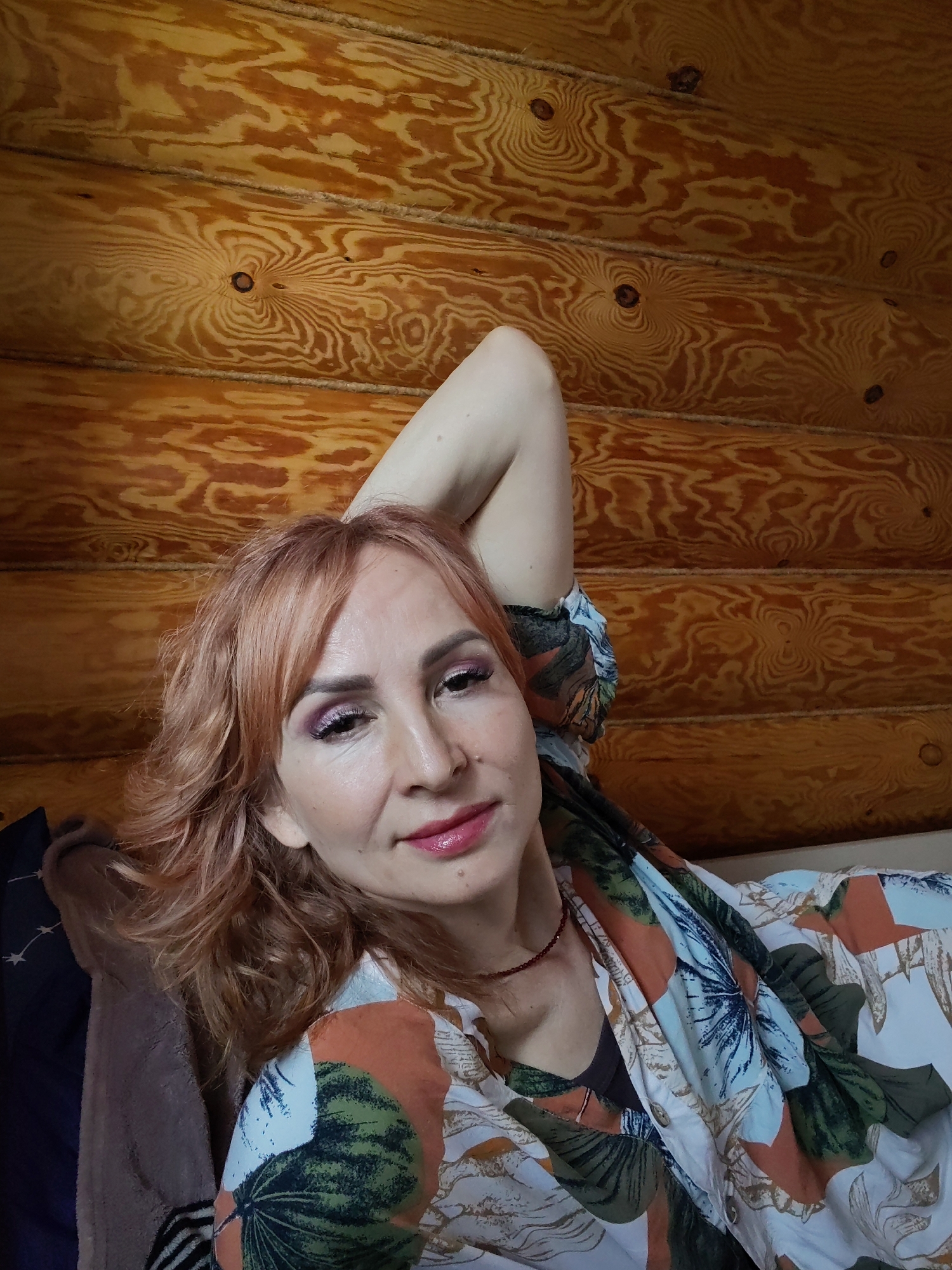 The image depicts a woman with reddish-blonde hair lying down in a log cabin in Старое Михайловское, Russia. She is wearing a colorful, patterned shirt and a red necklace. Her left arm is raised above her head. She is facing the camera with a slight smile. The background is comprised of stacked wooden logs forming the wall of the cabin.