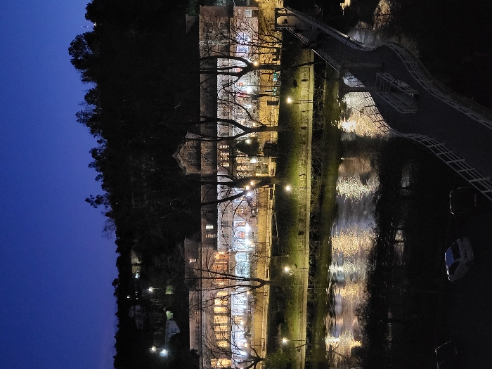Night scene depicts a building with multiple floors illuminated with lights. A river reflects these lights, creating a shimmering effect. A bridge extends over the river, connecting both sides. Bare trees are aligned along the river bank, interspersed with lampposts. The sky is a deep blue, transitioning into darkness towards the top. A hillside covered with trees forms the backdrop.