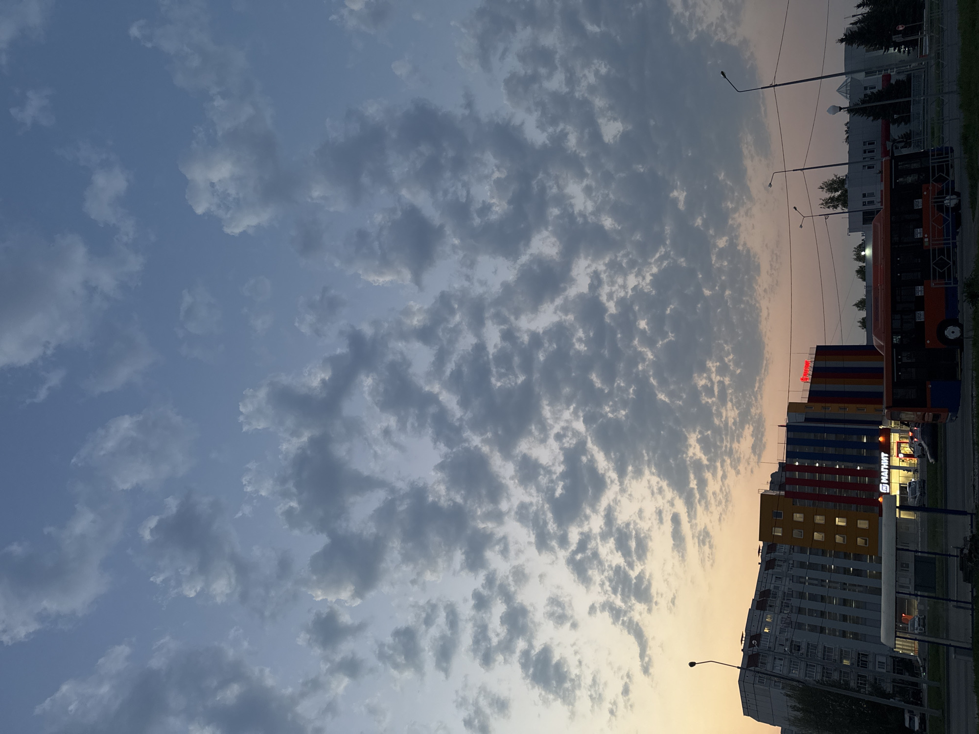 The image depicts a cityscape at twilight. The sky is filled with textured clouds, transitioning from a light blue to a pale orange near the horizon. In the lower right of the frame are several tall buildings, the tallest of which has colorful horizontal bands of yellow, red, and blue. A sign with the Cyrillic word "МАГНИТ" (Magnit) is visible, likely indicating a grocery store chain commonly found in Russia. A bus, colored orange and dark blue, is parked alongside the building. Streetlights line the street, and greenery is seen along the edges of the frame, suggesting trees or bushes. The location is Unknown, Unknown.