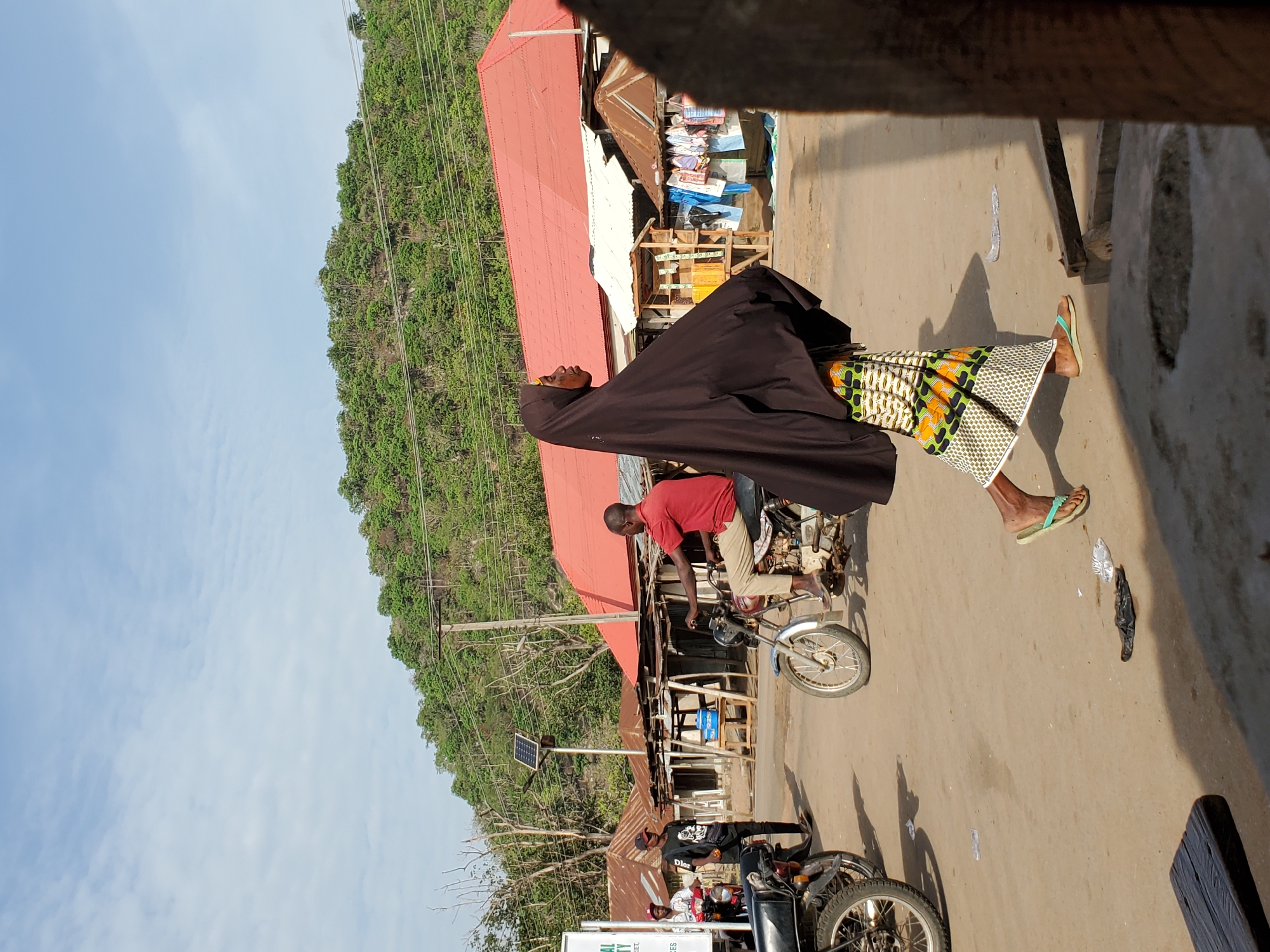 This photograph captures a bustling street scene in Mararaba, Nigeria, under bright, clear skies. The time of day appears to be midday or early afternoon, indicated by the strong, distinct shadows cast by people and objects on the dusty road.
In the foreground, slightly to the right, a woman stands or walks, adorned in a long, dark brown abaya or cloak that covers most of her body, revealing a vibrant, patterned skirt beneath. The skirt features geometric designs in green, orange, white, and black. She wears light green flip-flops. Her face is seen in profile, looking slightly upwards.
To her left, a man rides a motorcycle, wearing a red short-sleeved shirt and light-colored trousers. Further down the street, another man is partially visible on a parked motorcycle, wearing a black t-shirt with the word "Dior" faintly visible. Behind him, another person in a white and red patterned shirt can be seen.
The street is unpaved and dusty, with visible debris like plastic bags on the ground. Buildings with corrugated iron roofs, some appearing weathered and others newer, line the street. These structures likely house small shops or stalls, with various goods and items visible within them. A small solar panel is mounted on a pole near the parked motorcycles, suggesting a blend of traditional and emerging infrastructure.
The background is dominated by a large, verdant, tree-covered hill, emphasizing the natural landscape surrounding the settlement. Utility poles and power lines crisscross the scene, extending up the hillside. The image portrays a vibrant snapshot of daily life and movement in a semi-urban or rural Nigerian setting.