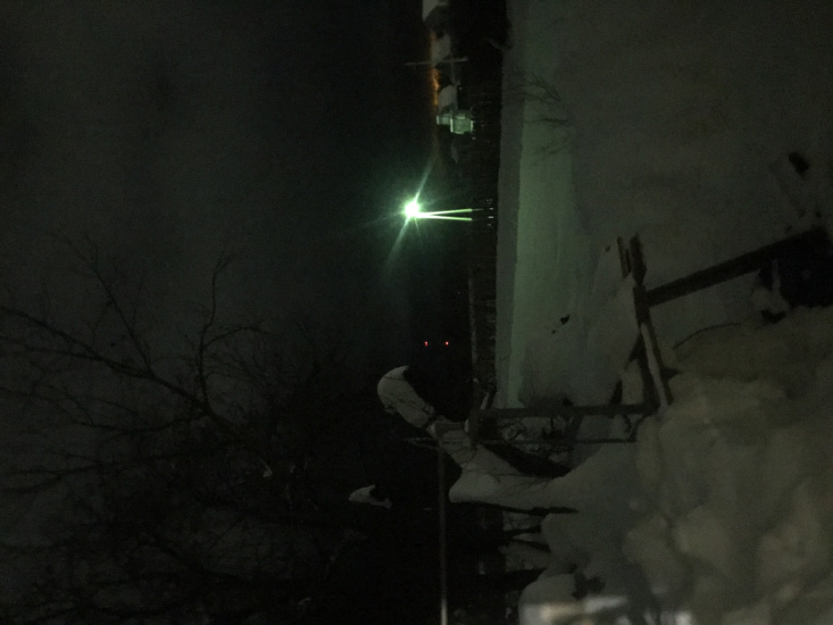 An outdoor night scene shows a white-covered wall with a wooden structure resembling steps. A bright light shines above, with two red spots appearing below it. Leafless trees stand to the left against the dark sky. Snow is visible on the wall and ground. The image is dimly lit, and the elements are primarily in shadow.