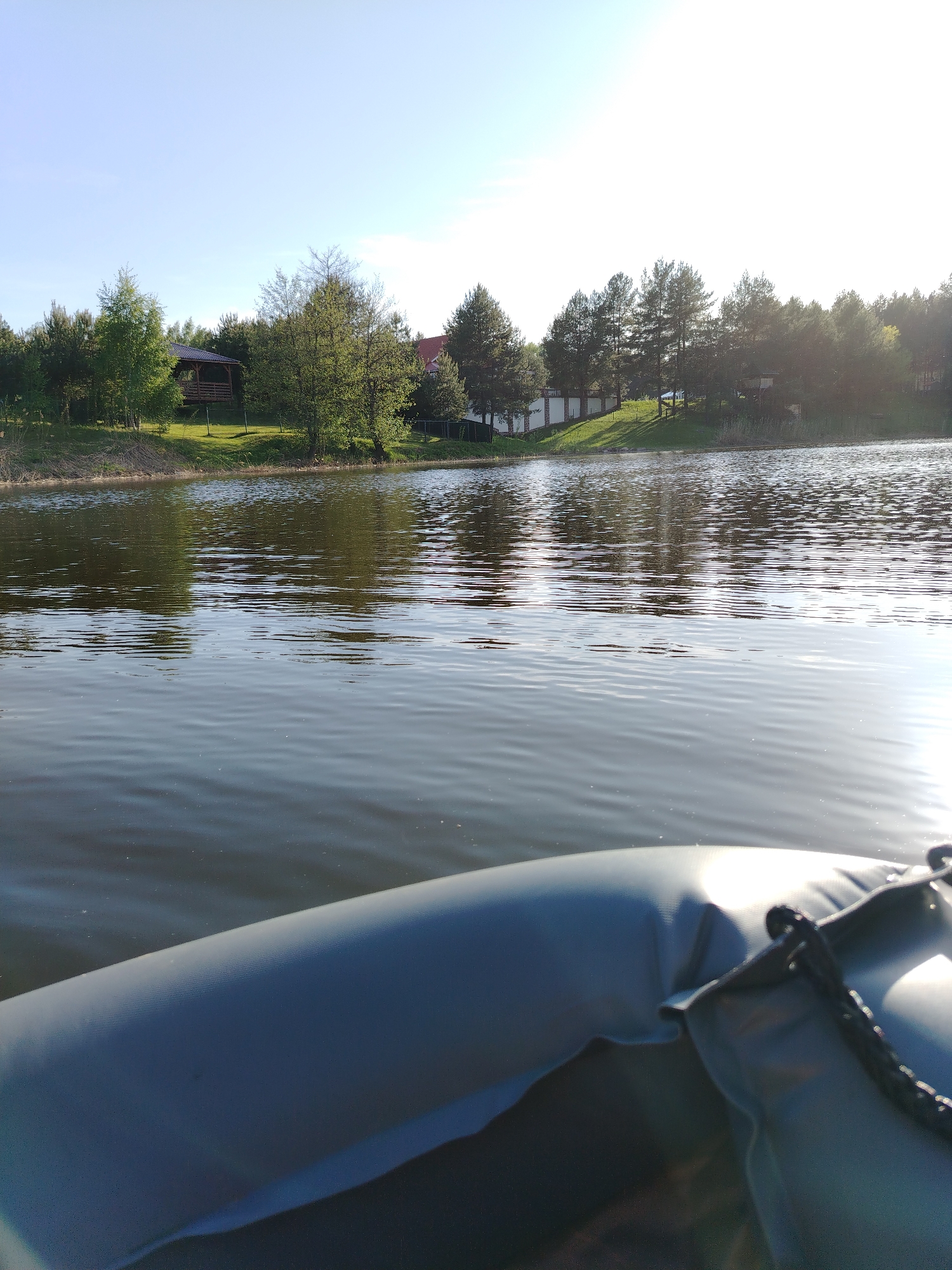 The photograph presents a view from a small boat on a lake. The hull of the boat is visible in the foreground. Beyond the boat, the lake reflects the light of the sky and the trees lining the opposite shore. On the shore, a structure resembling a gazebo is visible among the trees to the left, and houses are visible on the right behind a white fence. This scene is located in Stroitel', Russia.