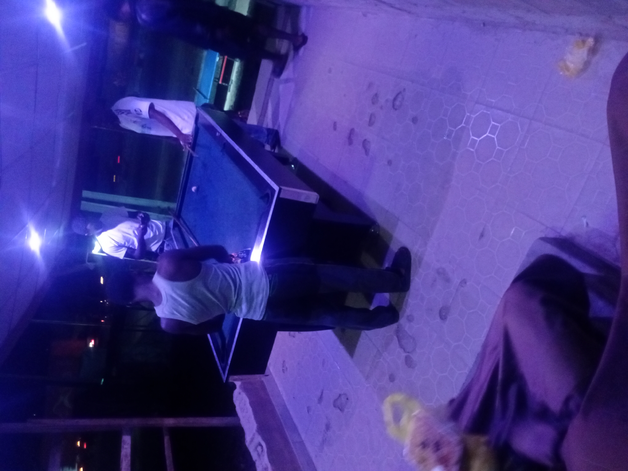 This image, taken at night in Lokoja, Nigeria, depicts a lively scene within what appears to be a recreational establishment, likely a bar or game room. The space is bathed in striking artificial purple and blue lighting, casting a cool, vibrant glow over the environment.
The central focus is a pool table with a blue felt surface, around which several individuals are gathered. One person, wearing a white tank top and dark pants, stands with their back to the camera, positioned next to the table, seemingly engaged in a game of billiards. Another individual in a white t-shirt is visible further down the table, extending their arm as if taking or preparing for a shot. A third person's lower body, dressed in dark clothing, is visible in the bottom right foreground, suggesting they are seated or relaxing nearby, with a small yellow object (possibly a bag or crumpled wrapper) near their feet. A fourth figure can be vaguely seen in the darker background.
The floor features light-colored tiles with a distinctive geometric pattern of interlocking hexagons and octagons. The overall atmosphere suggests a casual and social evening activity centered around the game of pool. No discernible text is visible in the image.