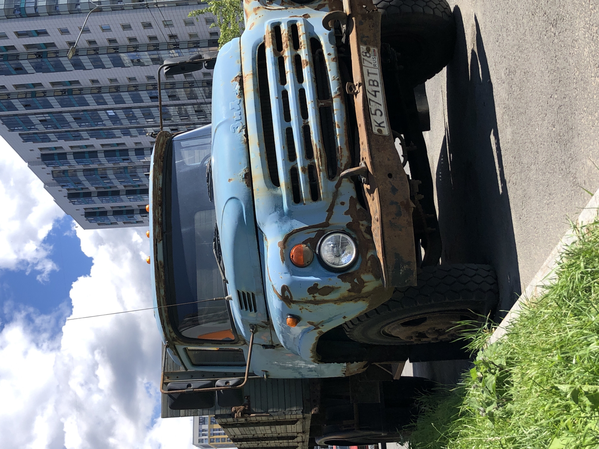 The image shows a rusty, light blue truck parked on the side of a paved road with a grassy verge. The truck is old and weathered, with significant rust on its hood, grille, and bumper. It has a round headlight and an orange turn signal on the front. A license plate is visible on the front of the truck, reading "K574BT 78 RUS". In the background, a modern, tall building with many windows is visible, suggesting an urban or suburban setting. The sky is blue with white, fluffy clouds, indicating it's daytime and likely fair weather. The scene is shot from a low angle, looking up at the truck and the building. The image is rotated 90 degrees clockwise.