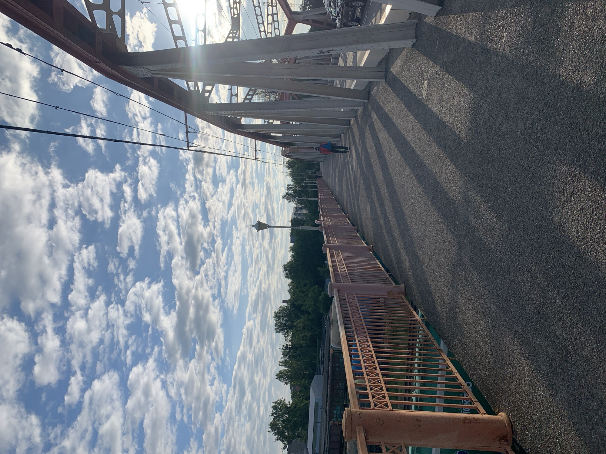 This is a daytime, eye-level, wide shot of a pedestrian walkway on a bridge. The walkway is made of asphalt and runs parallel to the side of the bridge. On the left side of the walkway is a red metal railing with decorative criss-cross patterns and vertical bars, supported by thick, round posts. Beyond the railing, there are train tracks and a train car. On the right side of the walkway are large concrete and red metal bridge supports, creating long, distinct shadows across the asphalt. The sky is blue with scattered, fluffy white clouds. In the background, there is a line of green trees and some buildings. A single person wearing a blue backpack and a red jacket is walking away from the camera on the walkway. To the far right, a dark-colored car is visible on a roadway alongside the bridge. The sun is shining from the upper right, casting lens flare.