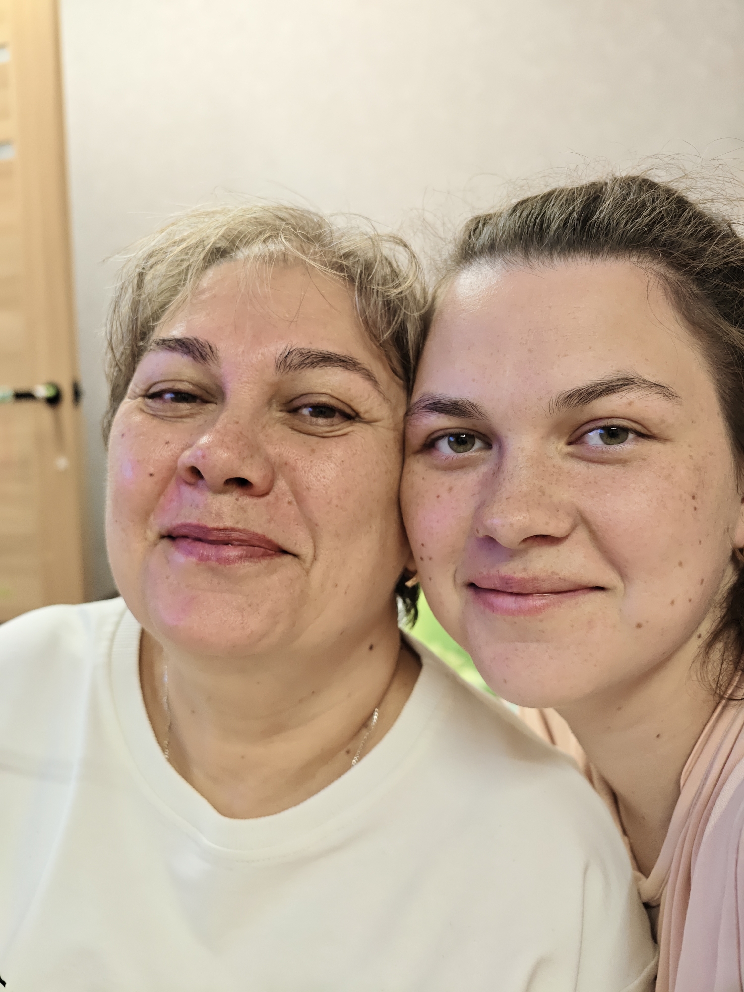 A close-up photograph depicts two Caucasian women, one older and one younger, posing cheek-to-cheek. The older woman, on the left, has short, light blonde hair, a round face, and is wearing a white V-neck shirt with a thin silver chain necklace. The younger woman, on the right, has dark hair pulled back, and freckles, and is wearing a light pink top. Both are smiling slightly at the camera. The background is a blurred mix of light-colored walls and a wooden doorframe, suggesting an indoor setting. The image was taken in Chaikovsky, Russia.