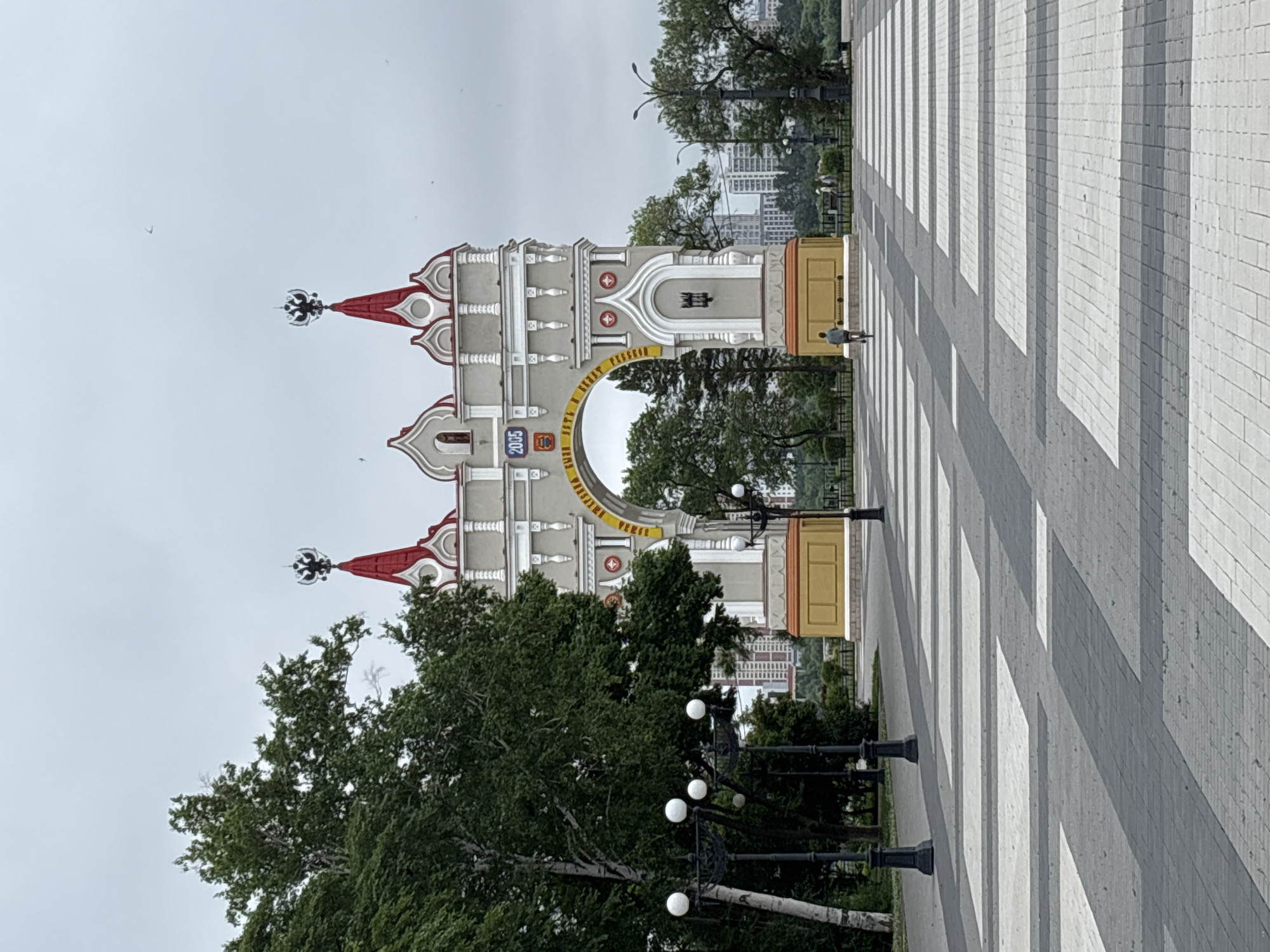 The image captures the Amur Triumph Arch in Blagoveshchensk, Russia, on a cloudy day. The arch is a prominent, ornate structure with a gray facade, red roofs topped with double-headed eagles, and yellow accents. Inscriptions are visible on the arch, and a digital display shows "2005."
In the foreground, a paved plaza with a checkered pattern of gray and white tiles leads towards the arch. Several lampposts with multiple white globes are positioned along the left side of the plaza. A single person is seen riding a bicycle across the plaza in front of the arch. Further in the background, trees line the perimeter of the plaza, and beyond them, several modern buildings and a body of water are visible, suggesting a riverside or waterfront location. The sky is overcast, contributing to the muted lighting of the scene.