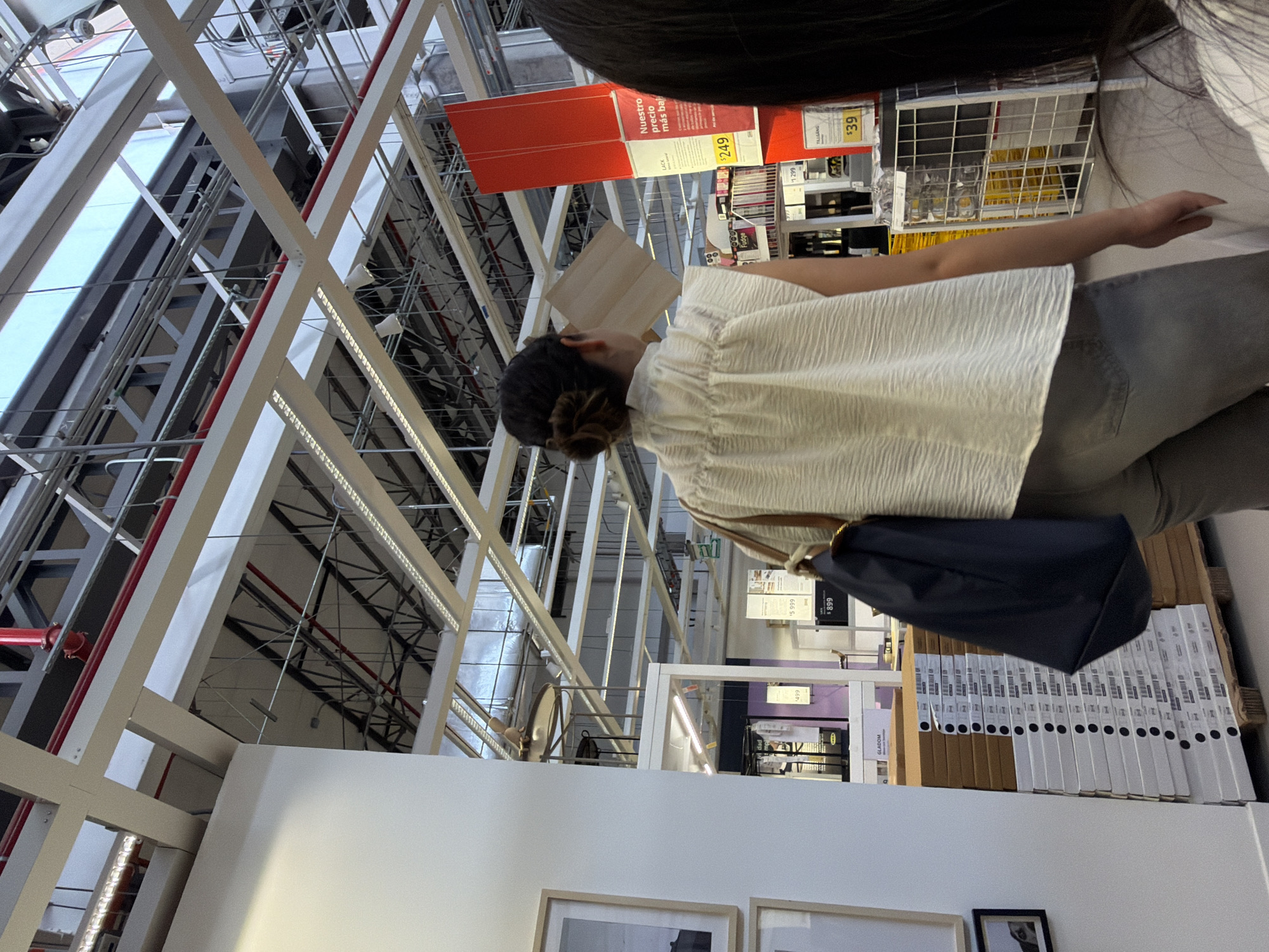 A person with dark hair pulled back in a bun, wearing a white top and gray pants, is seen browsing at an IKEA store. The person is standing next to shelves filled with products in boxes, with a dark blue handbag slung over their shoulder. Store signage, including price tags, is visible on the shelves. The store's infrastructure, including ceiling beams and lighting, can be seen in the background. The location is likely in a large retail space similar to IKEA, possibly in San Bernardino Tlaxcalancingo, Mexico.