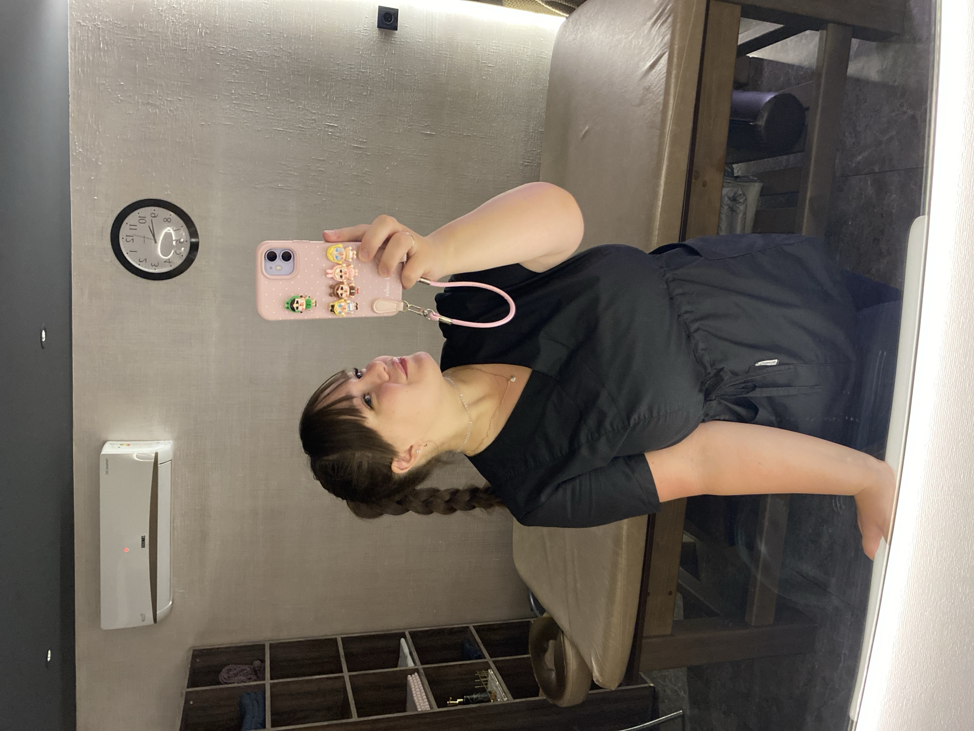 A woman with a braided ponytail and bangs, wearing a black scrub top and scrub pants, takes a mirror selfie in a dimly lit room. She is holding a pink phone adorned with cartoon figures. In the background, a beige massage table with a wooden frame is visible, along with a shelf unit containing folded towels and bottles. A clock on the wall shows approximately 3:08. An air conditioner is mounted on the wall above the shelf unit. The room appears to be a spa or treatment room. This scene is located in the city of Votkinsk, Russia.