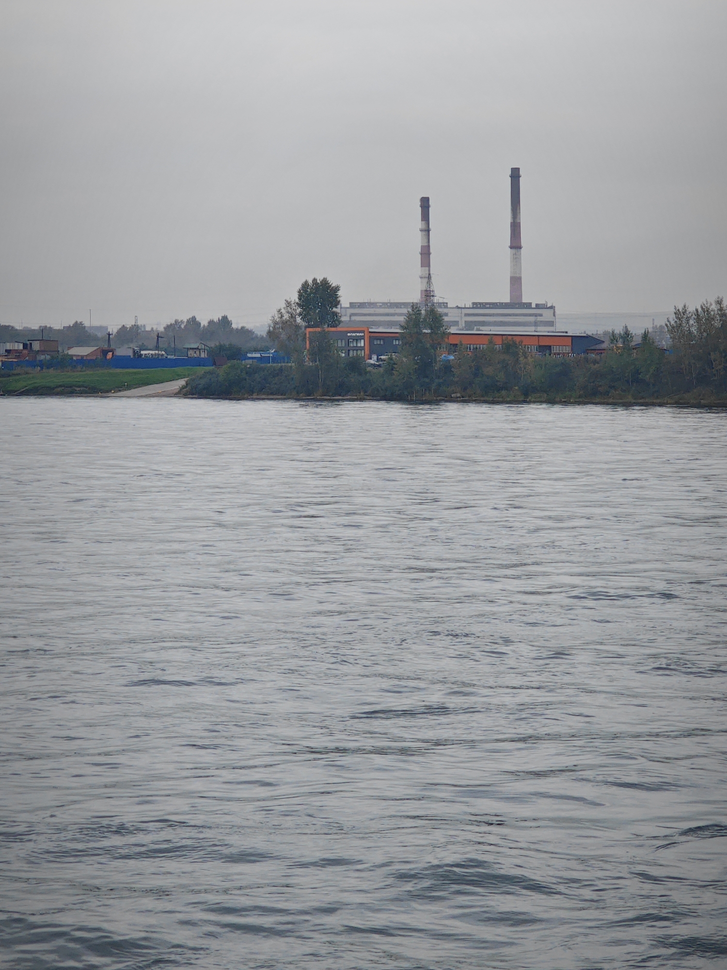 An industrial landscape is depicted from across a body of water. In the foreground, a wide, grey, rippling body of water occupies the majority of the frame, reflecting an overcast sky. The middle ground shows a shoreline with a strip of green grass leading to a ramp or pathway. Beyond the grass and a line of trees, several low-rise buildings with blue roofs and some industrial structures are visible. Dominating the background are two tall, cylindrical smokestacks, one significantly taller than the other, both with red and white banding. These smokestacks are part of a larger, grey industrial building complex. The sky is a uniform, pale grey, suggesting a cloudy or foggy day. The overall impression is one of an industrial area situated near a river or large body of water. The location is identified as Peschanovka, Russia. There are no people or animals visible in the scene.