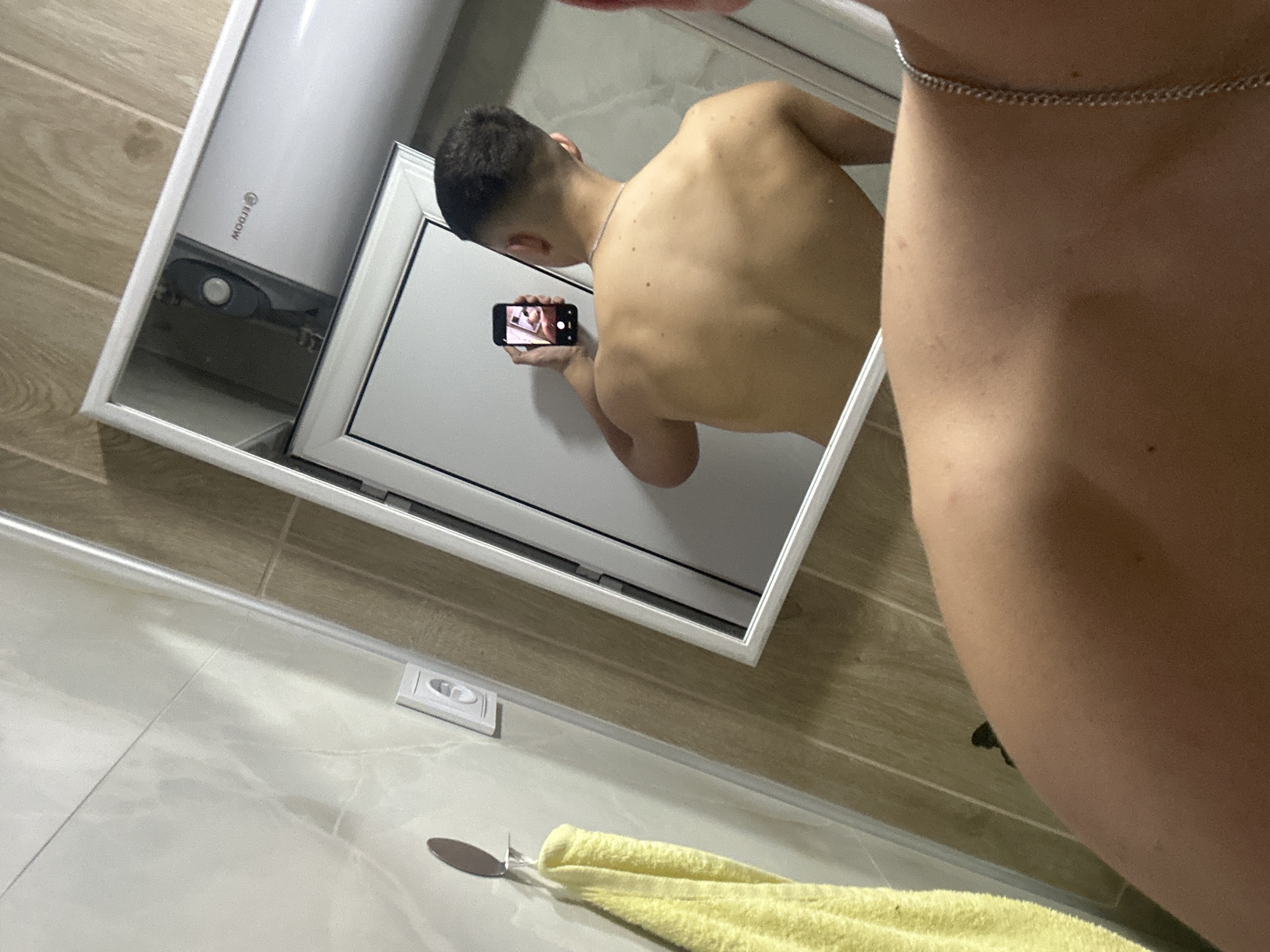 The image captures a mirror selfie taken by a person in what appears to be a bathroom. The primary view is a reflection in a rectangular mirror, framed in white, showing the back of a person from the waist up. The person has short, dark hair and is shirtless, revealing their bare back. They are holding a black smartphone with their right hand, positioned to capture the mirror reflection. On the phone screen, a smaller, recursive image of the same mirror reflection is visible. A thin silver chain is draped around the person's neck.
To the left within the mirror's reflection, a tall, white cylindrical object is visible, likely a water heater, with "MOGJI" (reversed) printed on its side and a circular dial or button at its base. Below this, a white rectangular panel, possibly a cabinet door or another part of the bathroom fixture, is reflected. The wall behind these objects has light brown or wooden-patterned tiles.
In the foreground, outside the mirror's frame, the person's right shoulder and upper arm are partially visible on the right side of the image, confirming they are the one taking the photo. A silver chain matching the one worn by the reflected person is visible around their neck/collarbone area. Below the mirror, a bright yellow towel is draped over an unseen object, possibly a towel bar or shelf. Further down, on a light-colored, possibly marble-patterned tiled floor or countertop, a white electrical outlet is visible. A small, dark, hook-like object is also present near the towel. The overall lighting suggests an indoor setting, likely artificial light.
