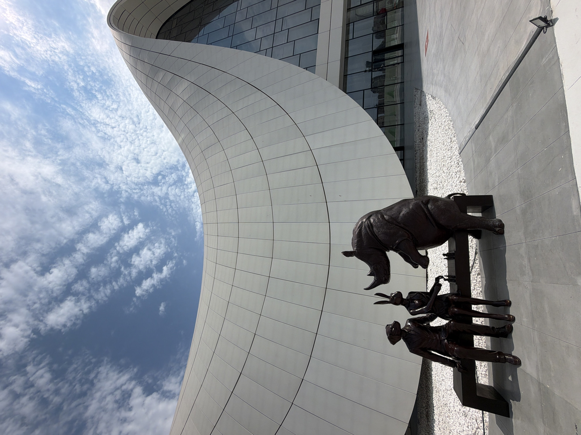 The image depicts a view of the Heydar Aliyev Center in Baku, Azerbaijan, on a bright, cloudy day. In the foreground, three bronze statues are arranged on a dark bench. From left to right, the statues represent a dog wearing a suit, a rabbit in a dress, and a rhinoceros. The dog and rabbit are seated, facing each other, with a chessboard between them. The rhinoceros is standing, leaning towards the chessboard. The statues are positioned in front of a white, curved building with a prominent, wave-like roofline. The building's facade is composed of numerous white panels. A large glass-paneled section of the building is visible on the right side of the image. The ground in front of the building is paved with grey tiles, and a curved area filled with white gravel is situated between the statues and the building. The sky above is blue with scattered white clouds. A single black pole with a small light fixture is visible on the pavement in the lower right corner.