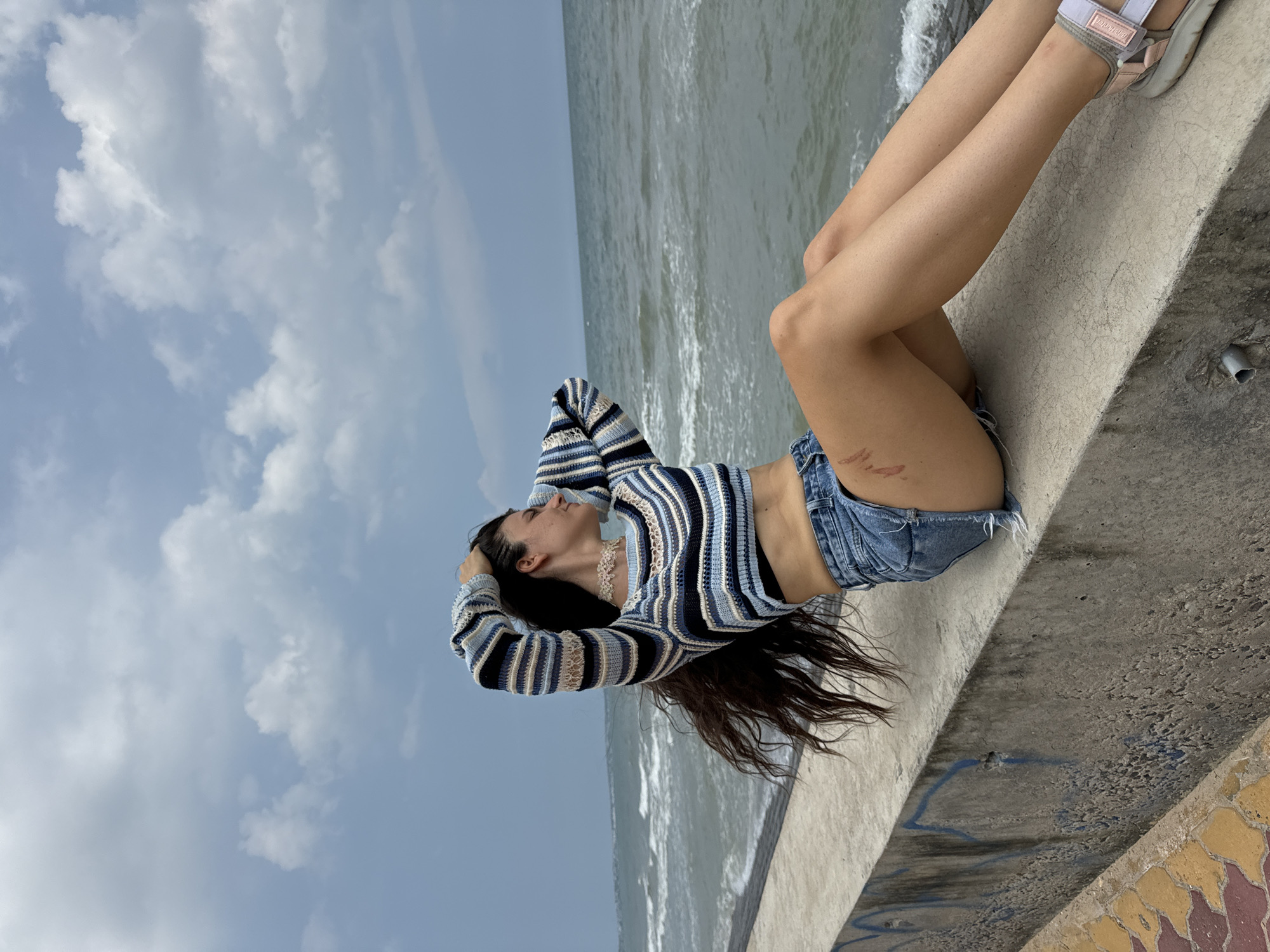A young woman with long dark hair is sitting on a concrete seawall, looking out at the ocean. She is wearing a striped crop top and denim shorts. Her arms are behind her head, and her hair is blowing in the wind. The ocean is rough, with waves crashing against the shore. The sky is cloudy, with a mix of blue sky and white clouds. The scene appears to be located in Mũi Né, Vietnam, a coastal town known for its beaches and sand dunes.