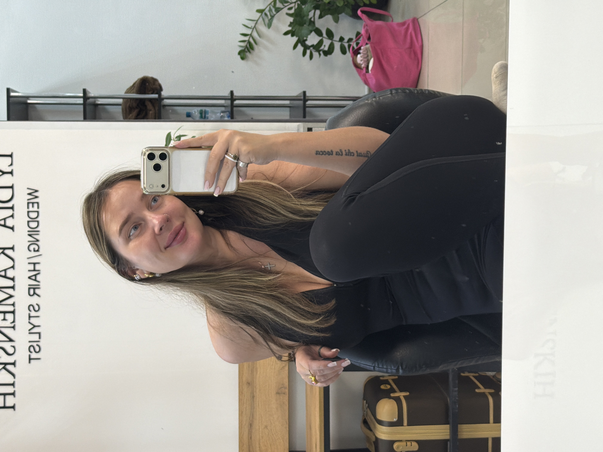 The image shows a woman with long blonde hair taking a selfie indoors. She is wearing a black top and leggings, seated in a black chair. A tattoo is visible on her arm. Her phone is a gold iPhone. Behind her are plants, a pink bag, and a rack of bottles. A piece of luggage is near the chair. A sign displaying the text "HIKIH KAMENEV HAIR STYLIST" is next to her. The location is the city of Chaikovsky, Russia.