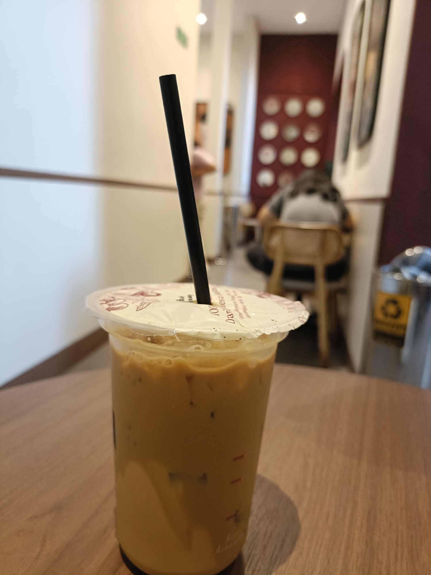 A close-up, low-angle shot showcases a plastic cup of iced coffee, its amber liquid filled with ice cubes and some dark specks, topped with a sealed plastic lid adorned with floral patterns and text. A black straw juts out from the center of the lid. The cup rests on a wooden table, its surface reflecting the ambient light. In the background, a person is seated at a wooden chair, their back to the camera, wearing a grey top. Beyond them, a burgundy wall is decorated with a grid of circular plates. To the right, a silver trash bin with a yellow recycling symbol is visible. The scene appears to be set in a cafe in the city of Semarang, Indonesia.