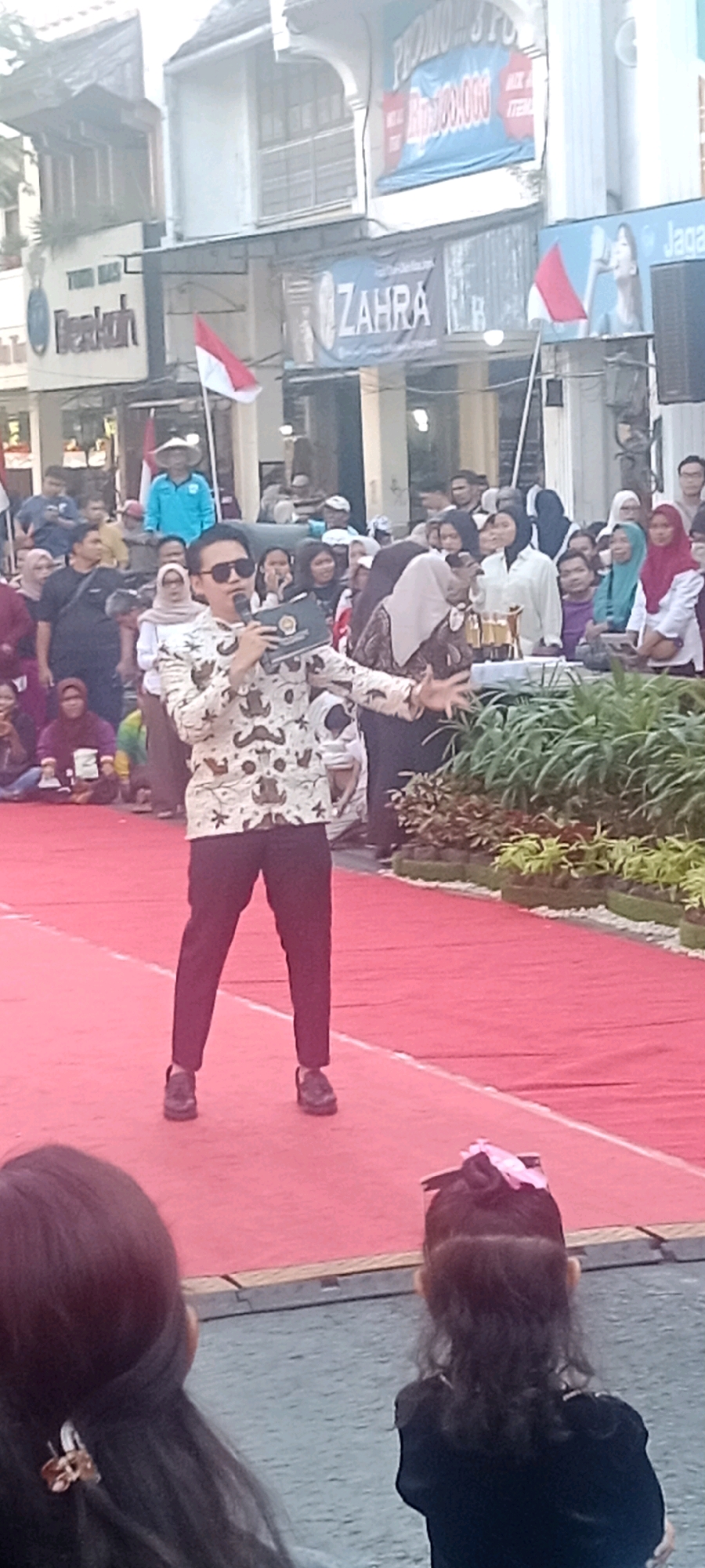 In Yogyakarta, Indonesia, a man in sunglasses and a patterned shirt stands on a red carpet, holding a microphone and a green book while speaking. He is surrounded by a crowd of people, many of whom are wearing headscarves. In the background, buildings are visible, including one with a banner that reads "ZAHRA." Indonesian flags are also displayed. The scene appears to be an outdoor event, possibly a festival or ceremony, given the red carpet and the festive atmosphere suggested by the crowd and the decorations.