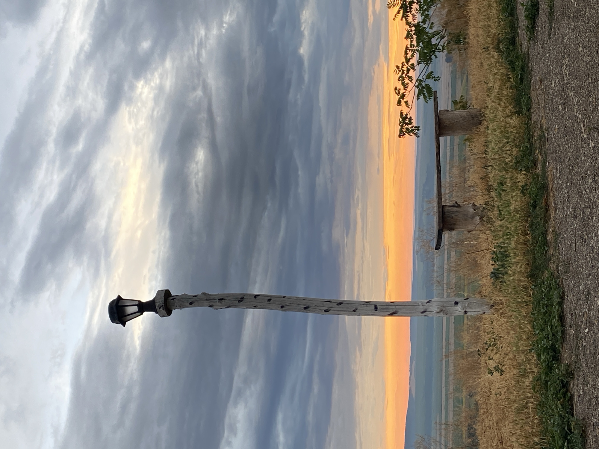 The image depicts an outdoor scene at dusk with a dramatic sky filled with gray and white clouds illuminated by an orange sunset glow on the horizon. In the foreground, a rustic wooden lamppost stands tall on a grassy knoll. To the right of the lamppost is a simple wooden bench, also appearing weathered, supported by log-like structures. The bench faces away from the viewer, overlooking a vast, rural landscape with rolling hills and fields that fade into the distance. The vegetation around the bench and lamppost consists of dry grasses and some green ground cover. The overall impression is one of a quiet, natural setting, possibly a viewpoint or resting spot, in the countryside of Horești, Moldova. There are no people visible in the image.